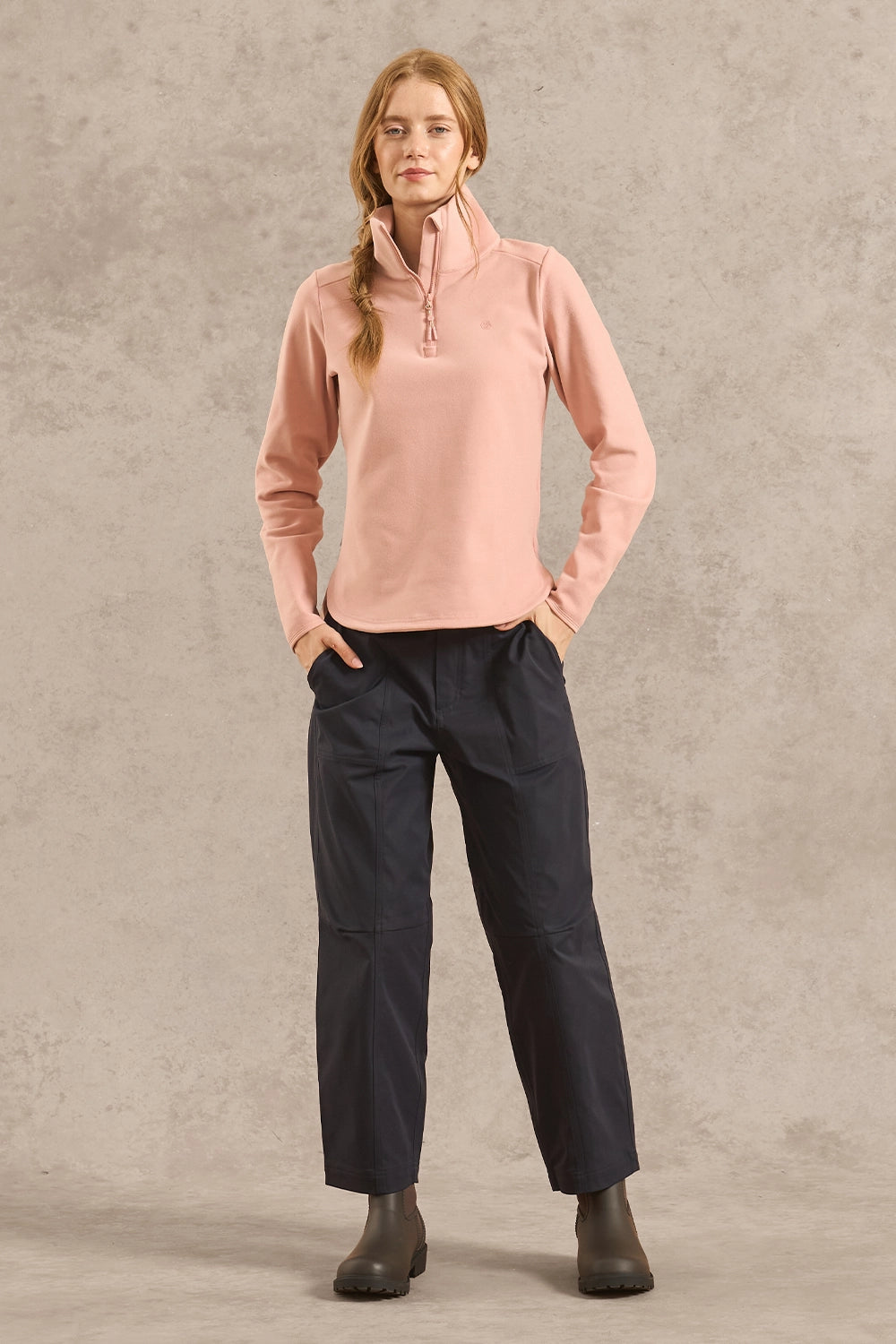 A woman modeling a light pink quarter-zip fleece pullover and dark navy pants, standing against a textured gray studio backdrop.
