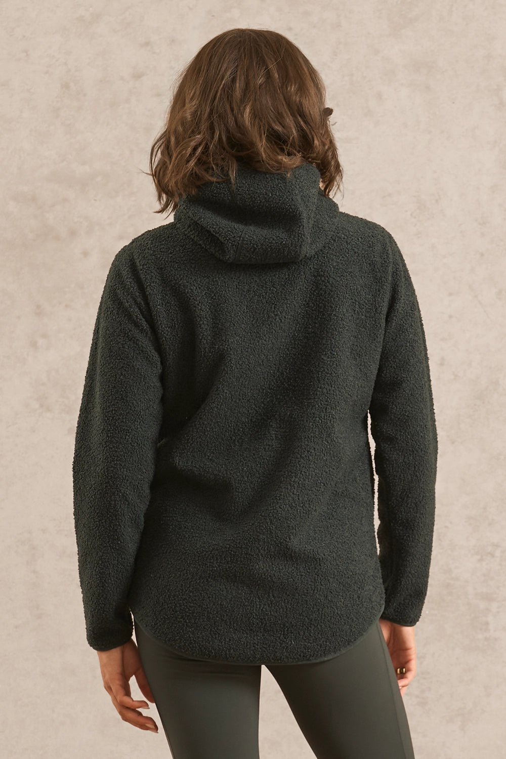A woman viewed from the back wearing a forest green sherpa fleece pullover hoodie and matching gray leggings, standing against a textured beige wall.