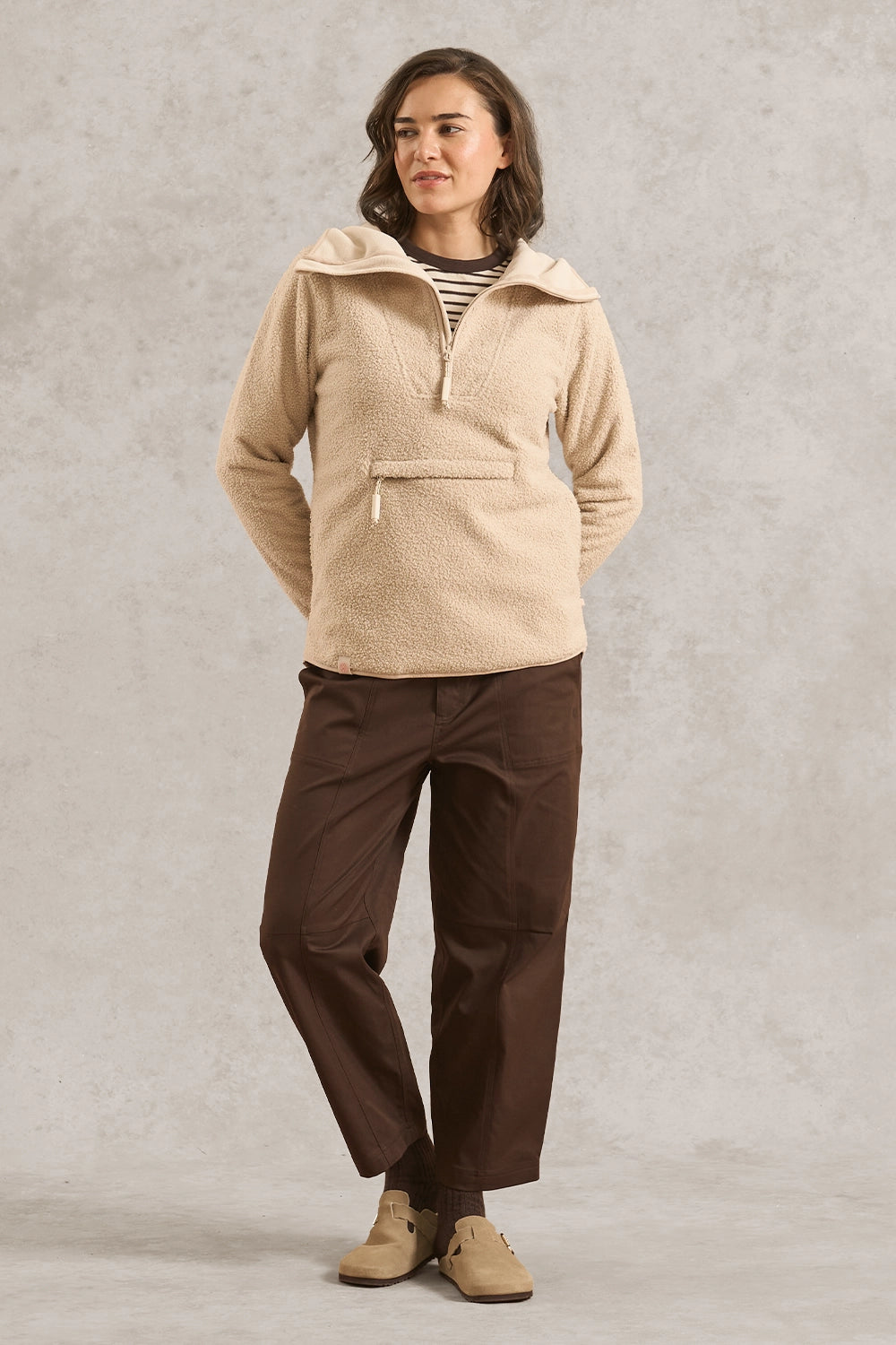A woman modeling a beige sherpa pullover with a half-zip and wide collar, paired with dark brown pants and tan shoes, standing against a textured gray studio backdrop.