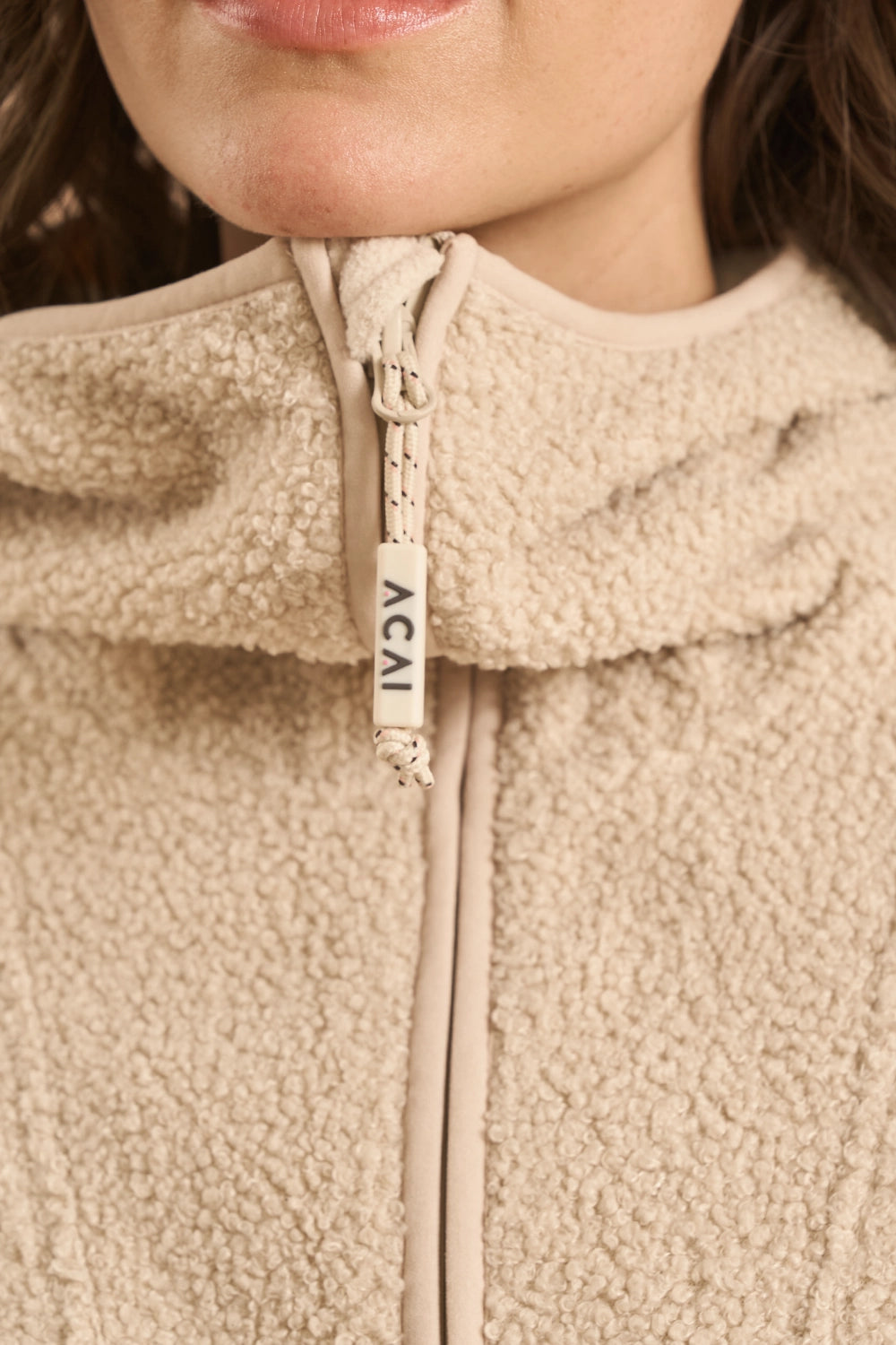 Close-up of a beige sherpa fleece zip-up jacket with a high collar and a cream zipper pull tab printed with the brand name 'ACAI'.