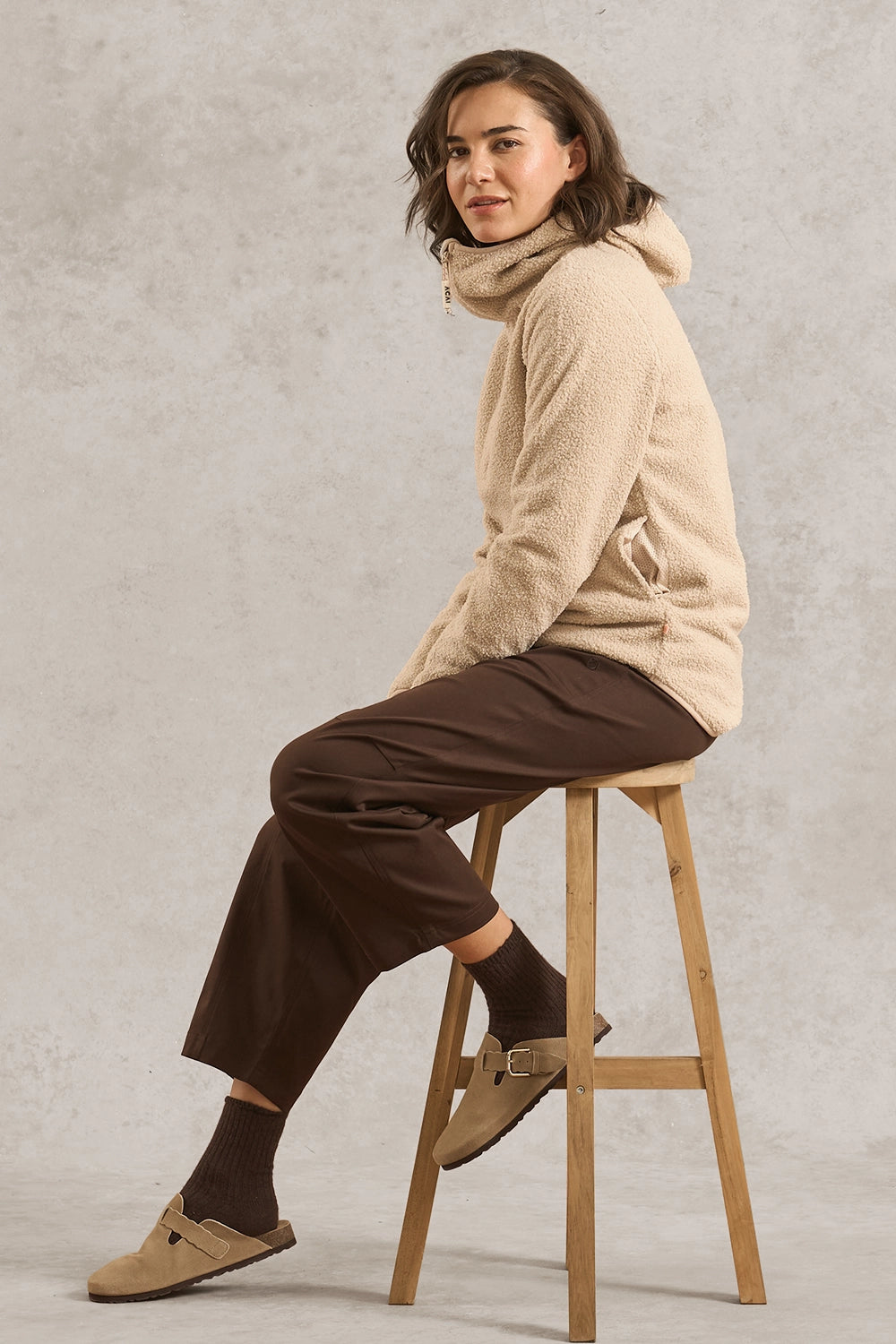 A woman sits on a wooden stool wearing a beige sherpa hoodie with a high neck, dark brown wide-leg pants, dark socks, and tan mule loafers against a textured gray studio background.