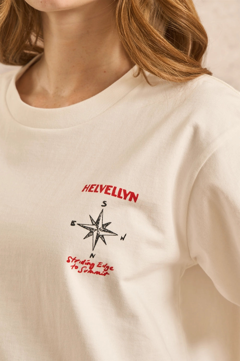 Close-up of a person wearing a cream-colored crewneck t-shirt with a red and black compass graphic and the text 'HELVELLYN' on the chest.
