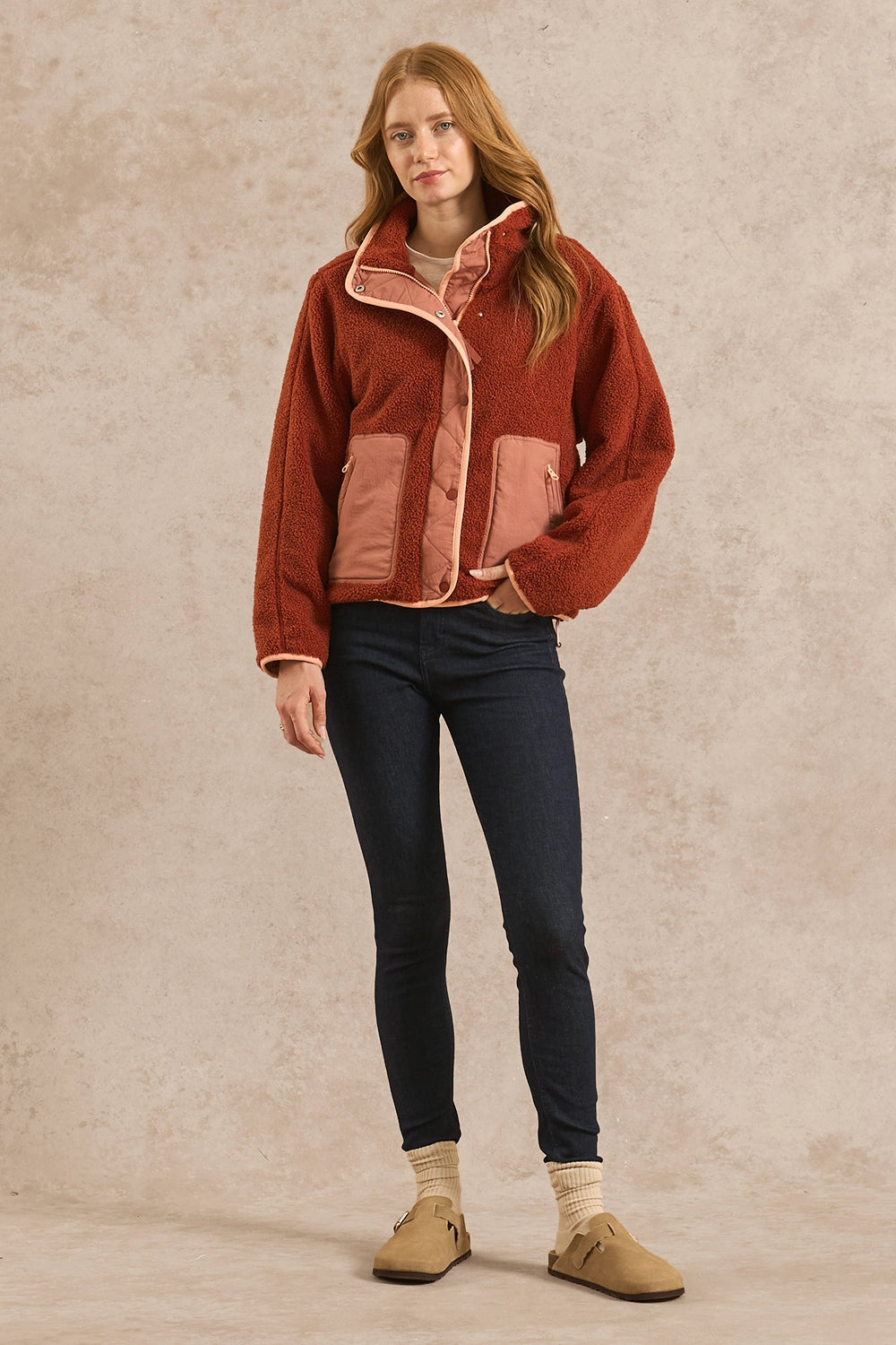 A woman stands against a textured beige studio backdrop wearing a rust-orange sherpa jacket with dusty pink lining, dark skinny jeans, cream socks, and tan slip-on shoes.