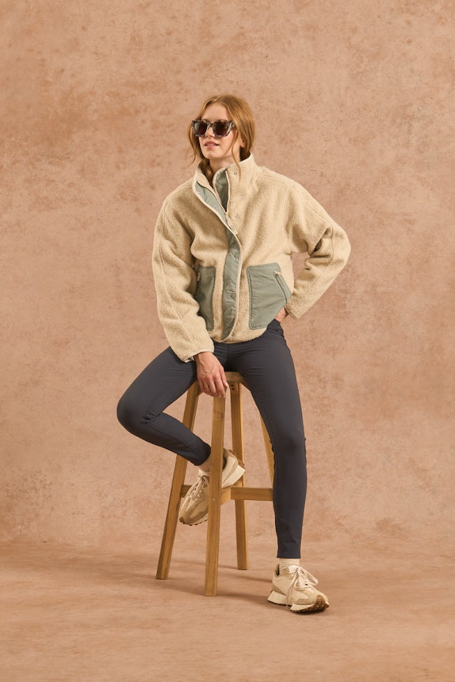 A model wearing a beige sherpa jacket with green and blue color-blocked panels, dark gray leggings, and beige sneakers, sitting on a wooden stool against a textured beige backdrop.