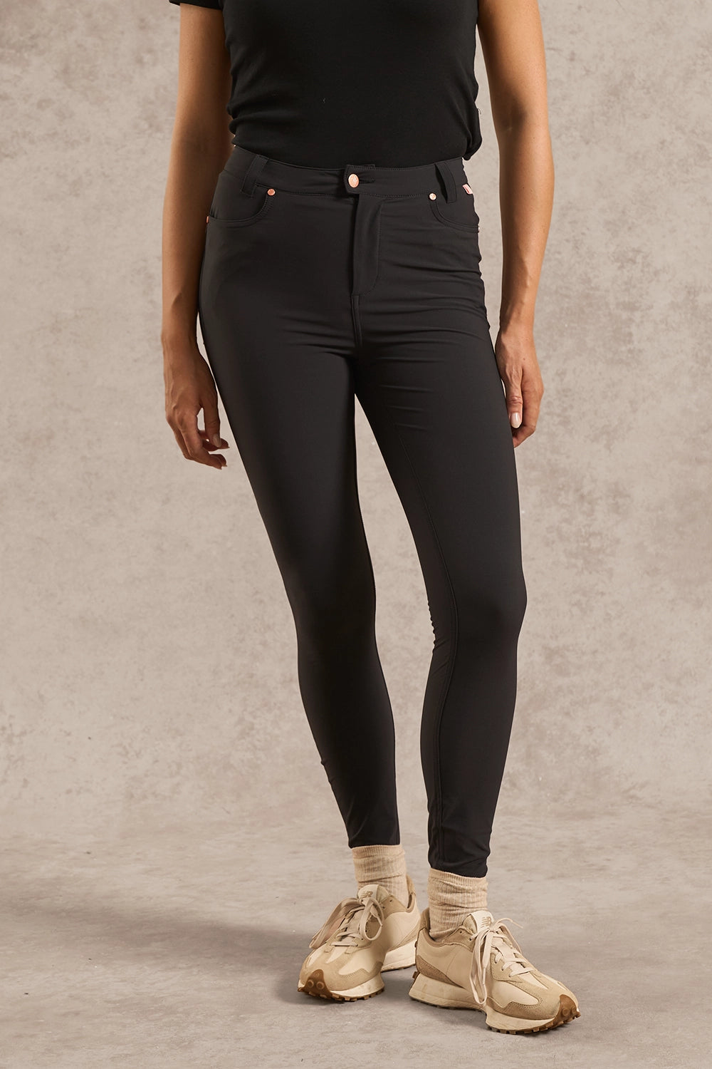 A model wearing black high-waisted skinny jeans, a black t-shirt, beige socks, and beige sneakers, standing against a textured gray studio backdrop.
