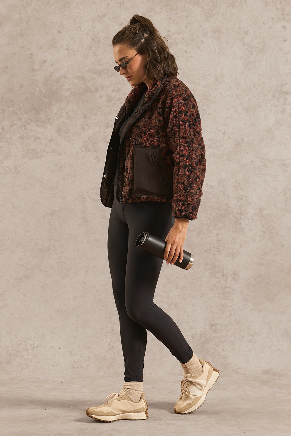 A woman wearing a dark brown and black patterned jacket, black leggings, beige sneakers, and sunglasses walks in profile while holding a black tumbler against a textured beige studio backdrop.