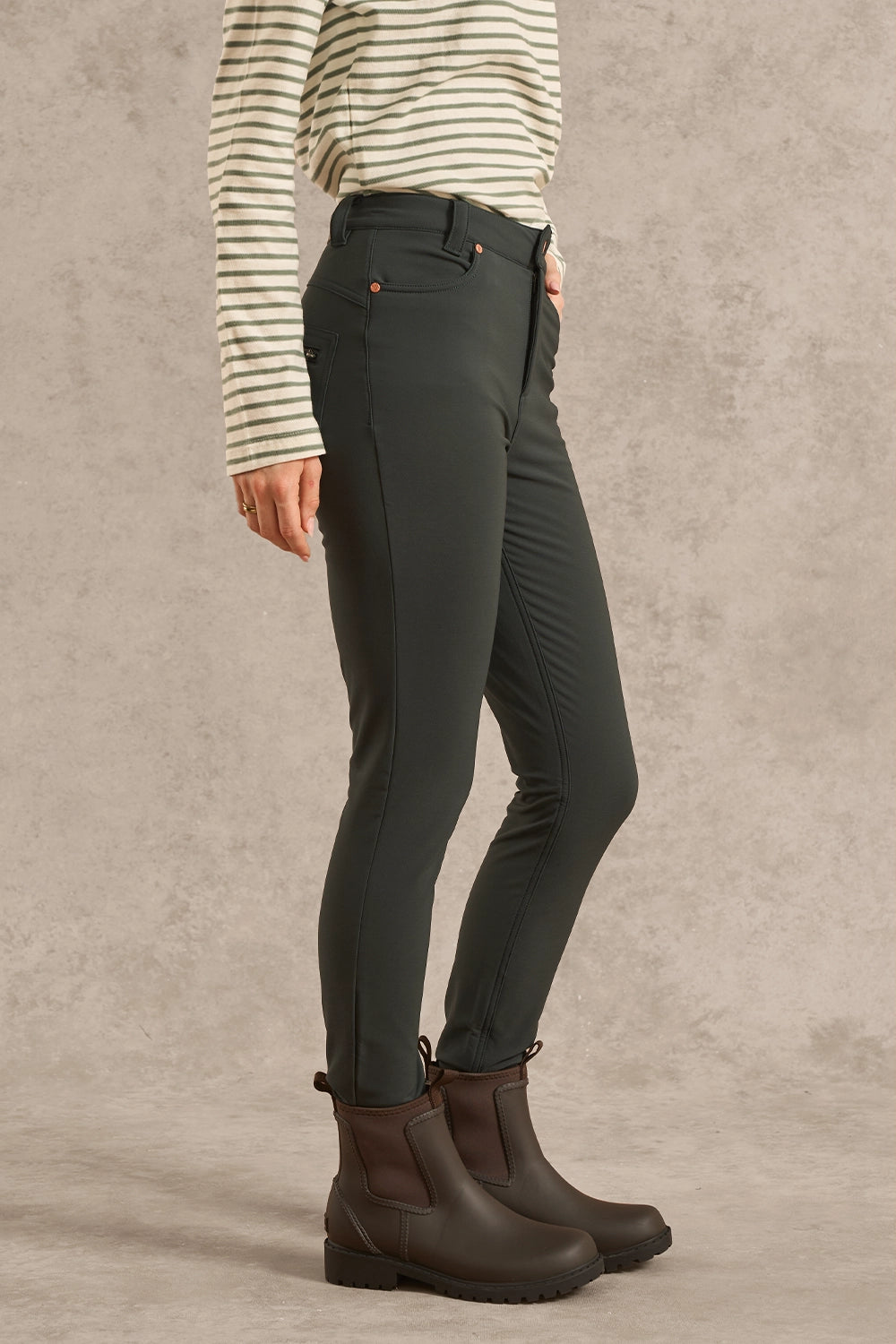 A model wearing dark olive green slim-fit riding breeches, a cream and green striped long-sleeve shirt, and dark brown Chelsea boots, shown from the waist down against a textured gray studio backdrop.