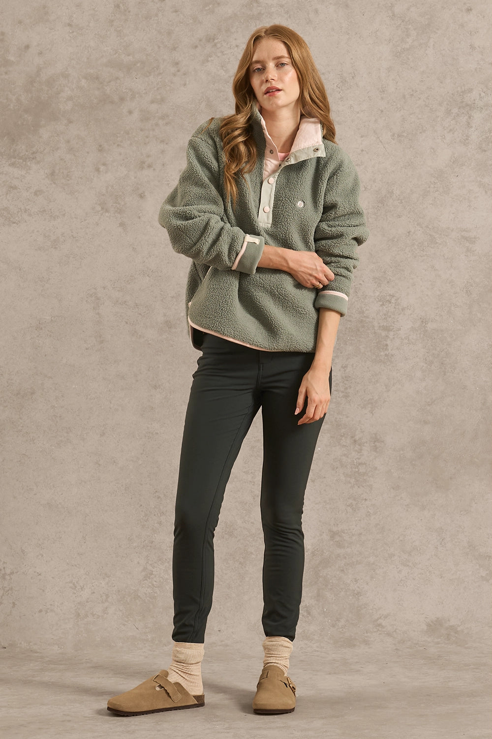 A woman stands against a textured gray studio wall wearing a sage green fleece quarter-zip pullover with cream sherpa lining, dark charcoal skinny pants, beige socks, and tan slip-on mules.