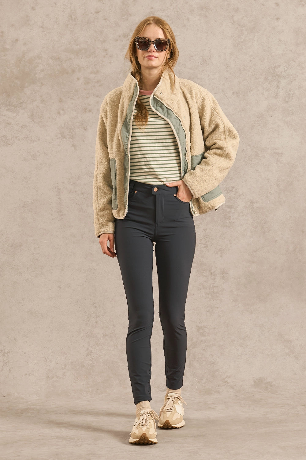 A woman modeling a beige sherpa-lined jacket over a green and white striped shirt, paired with dark gray skinny jeans and beige sneakers, against a textured beige studio backdrop.