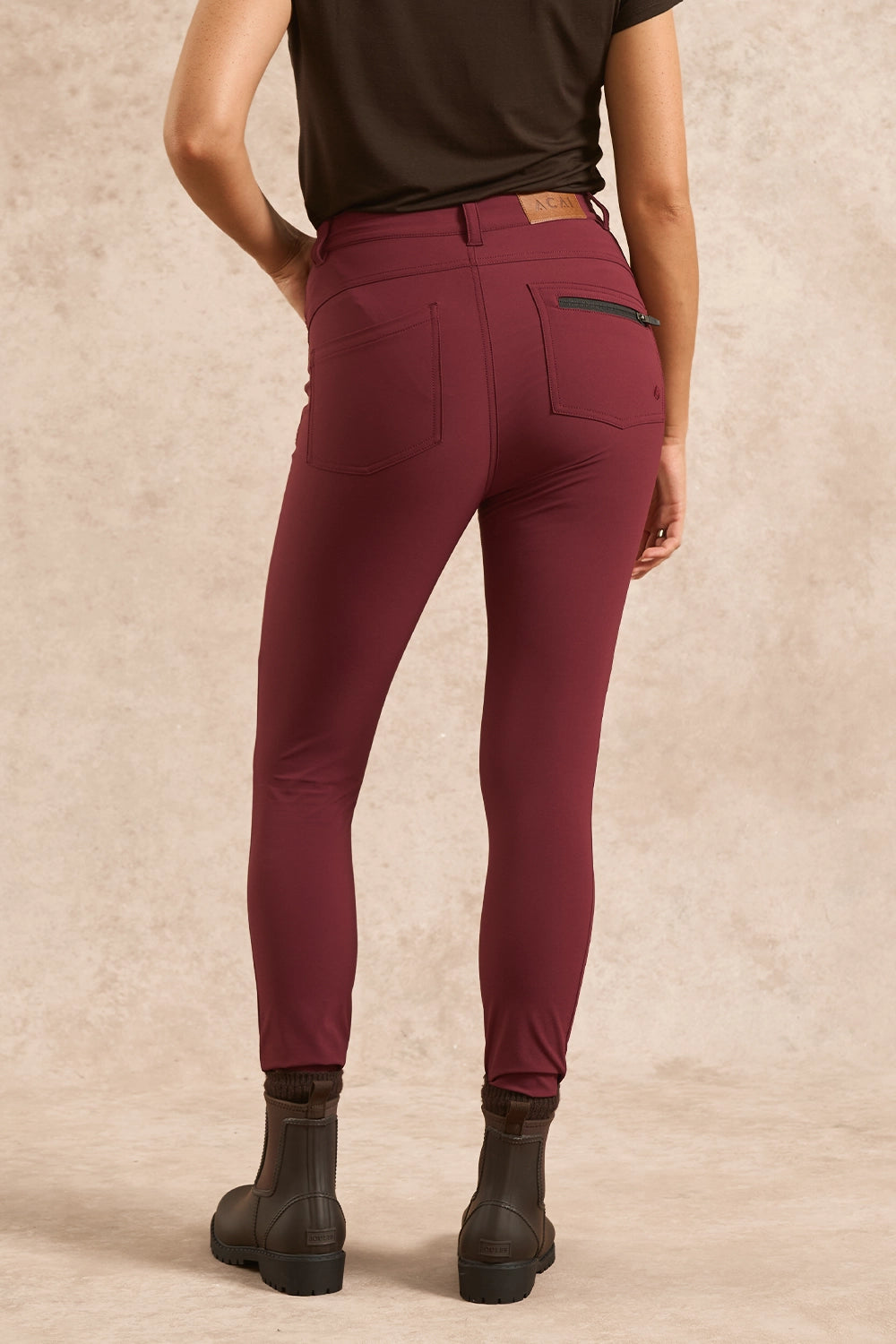 Rear view of a person wearing slim-fit burgundy riding breeches with a zippered pocket on the back right, paired with a dark brown top and dark brown boots, against a textured beige studio backdrop.