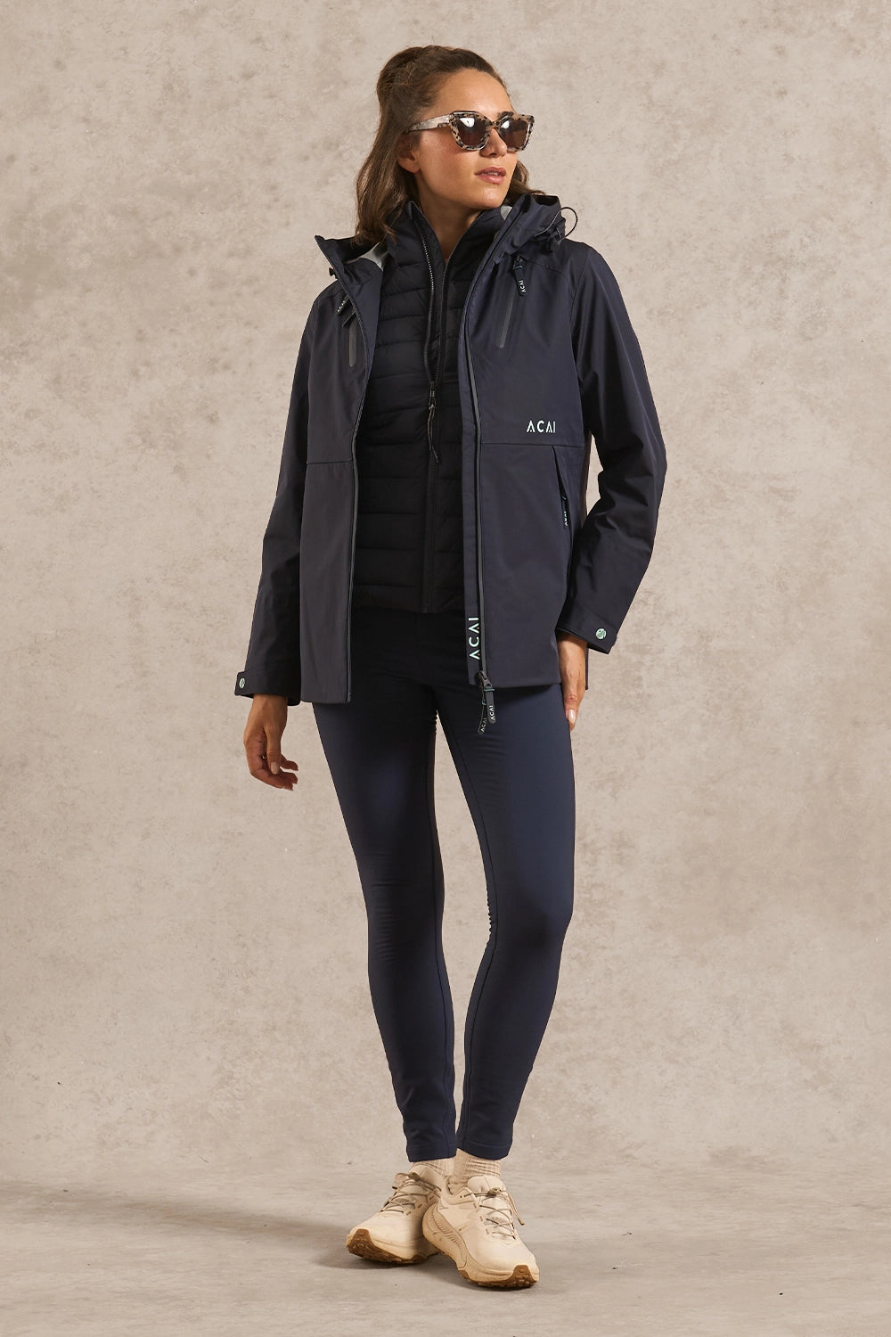 A woman modeling a dark navy outdoor jacket and matching leggings against a textured beige studio backdrop. The jacket has a small white ACHU logo on the left chest.