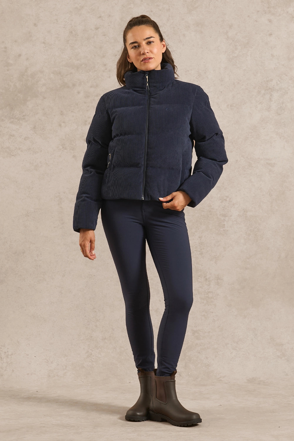 A woman modeling a navy blue puffer jacket, matching navy leggings, and dark brown rain boots against a textured beige studio backdrop.