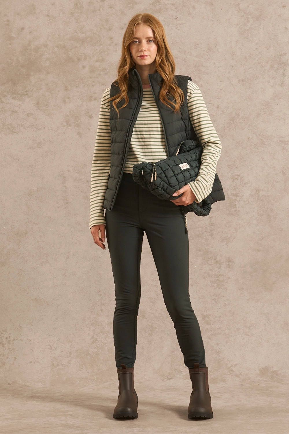 A woman stands against a textured beige studio backdrop wearing a dark quilted puffer vest over a cream and olive striped long-sleeve top, dark skinny pants, and dark brown ankle boots. She is holding a matching dark quilted crossbody bag.