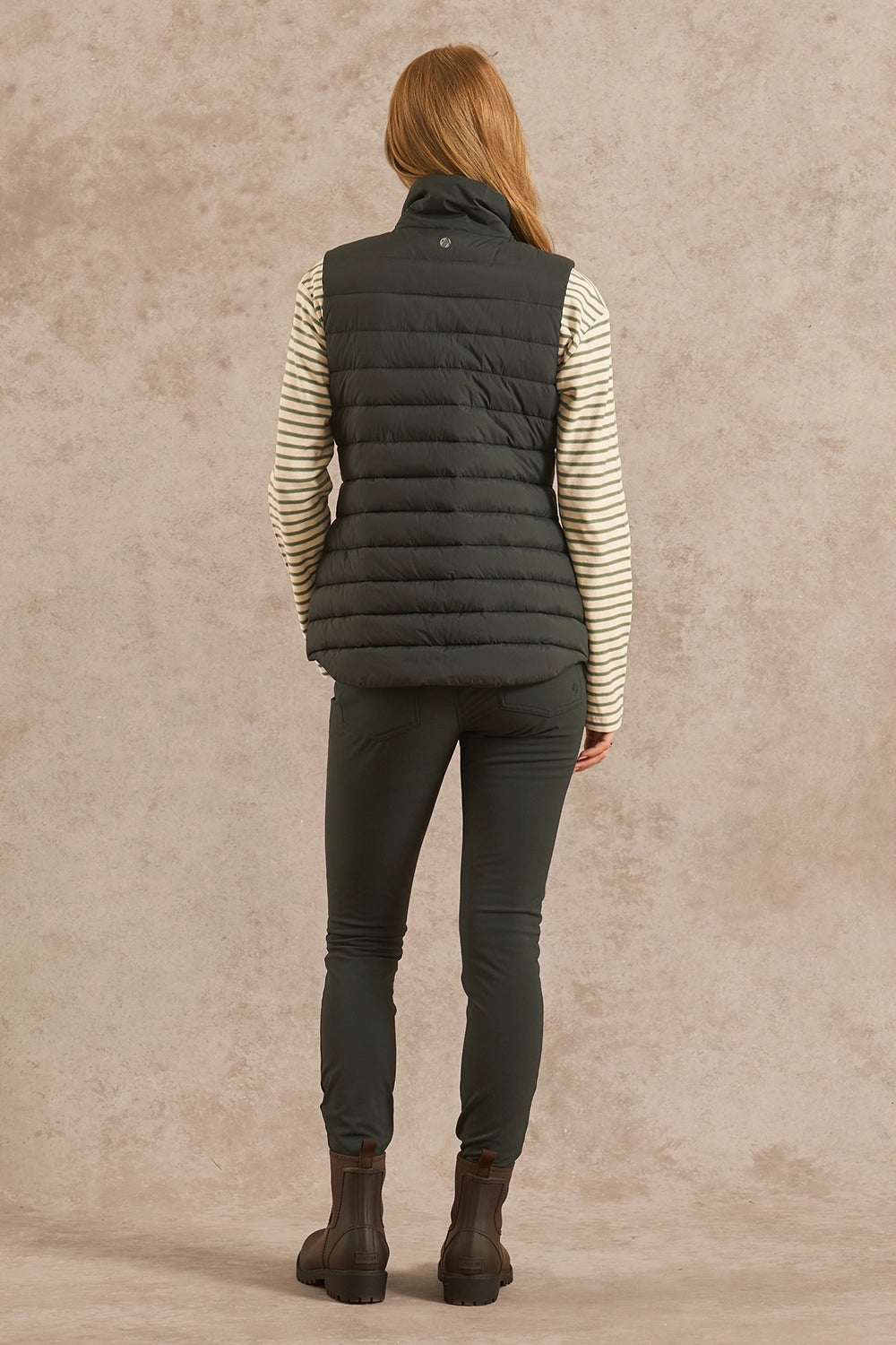 A woman is shown from behind wearing a dark olive green puffer vest over a cream and dark green striped long-sleeve shirt, dark green pants, and brown boots, standing against a textured beige studio backdrop.