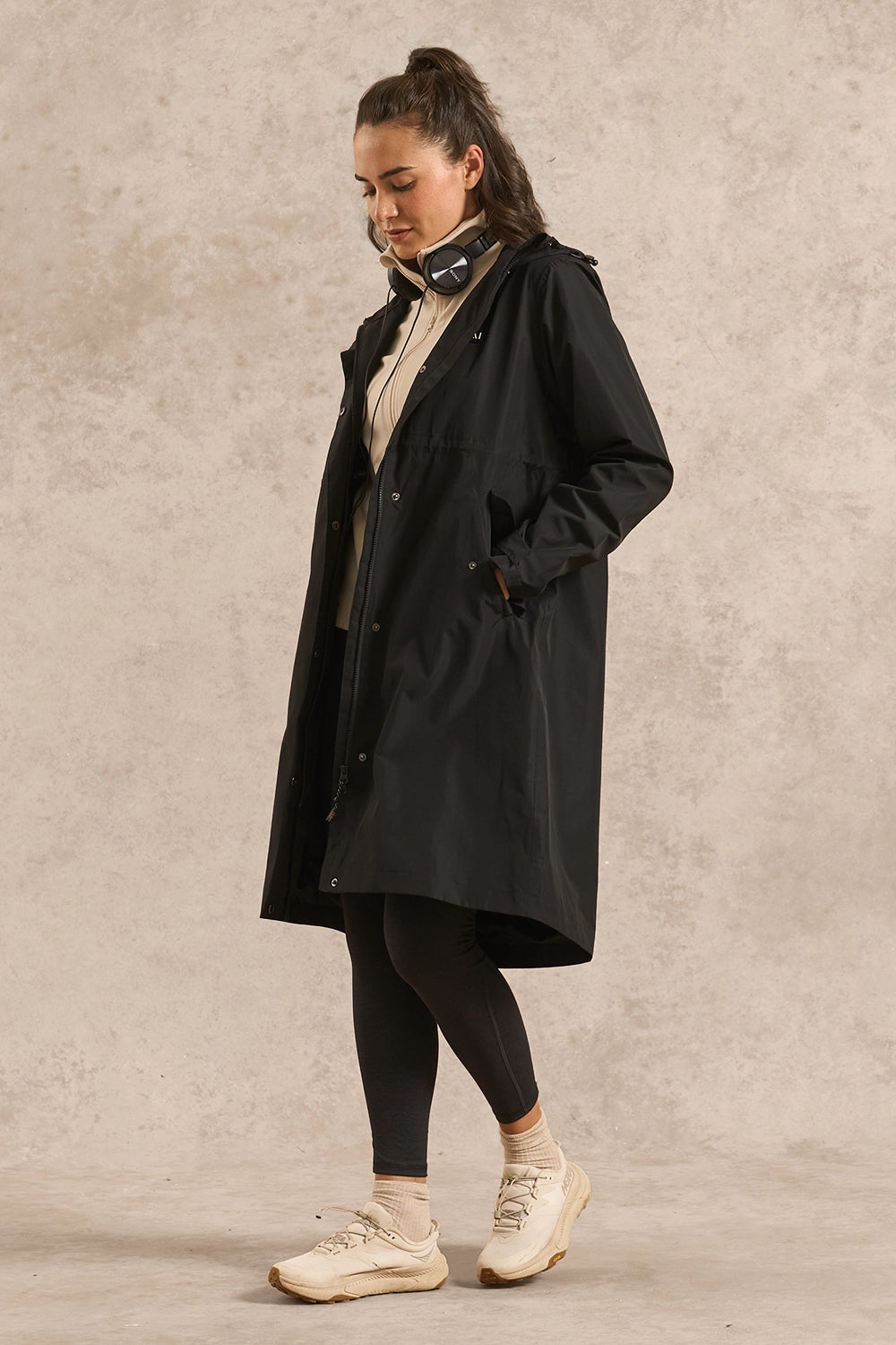 A woman wearing a long black hooded raincoat over a beige top, black leggings, and cream sneakers, standing against a textured light beige studio backdrop.
