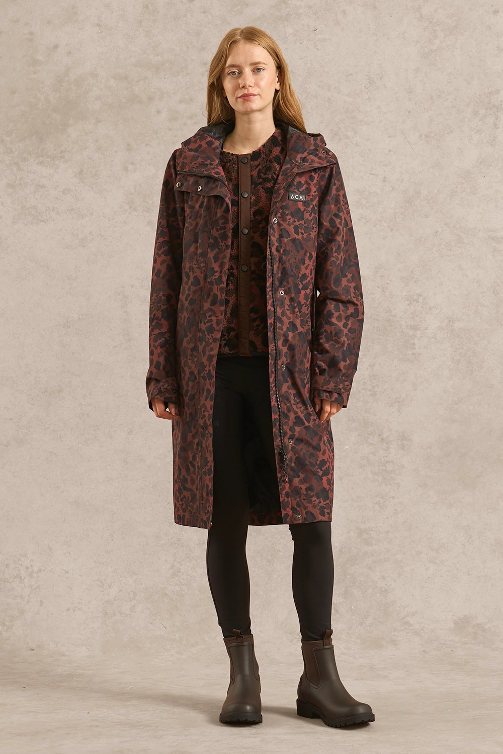 A woman stands against a textured light gray studio backdrop wearing a knee-length hooded coat with an allover dark red and black animal-style print, layered over a dark top, black skirt or pants, and dark ankle boots.