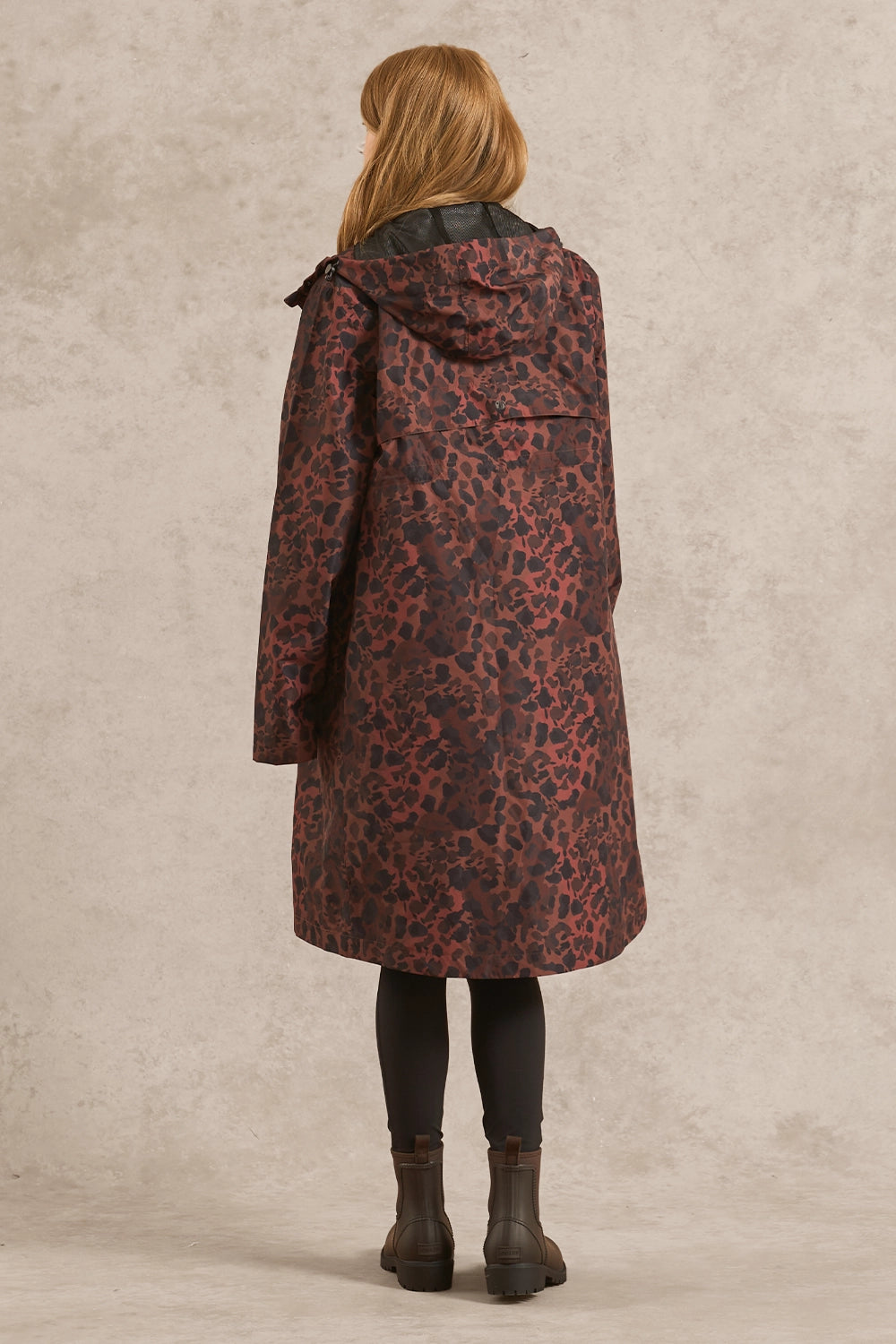 A woman viewed from the back wearing a hooded coat with a dark red and black leopard print pattern, paired with black leggings and dark brown boots, standing against a textured light gray studio backdrop.