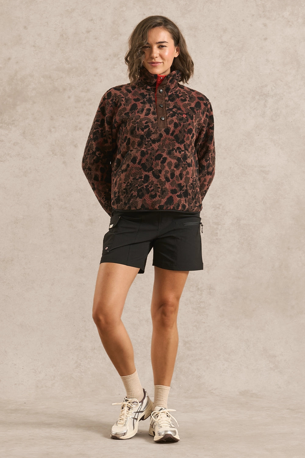 A woman modeling a brown and black leopard-print quarter-zip fleece pullover, paired with black athletic shorts, beige socks, and white sneakers, standing against a textured beige studio backdrop.