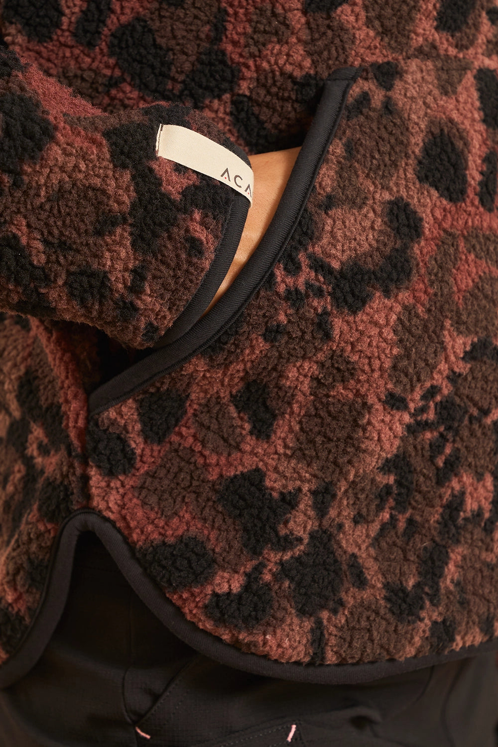Close-up of a textured fleece jacket with a brown and black leopard print pattern, featuring black trim and a visible cream brand label.