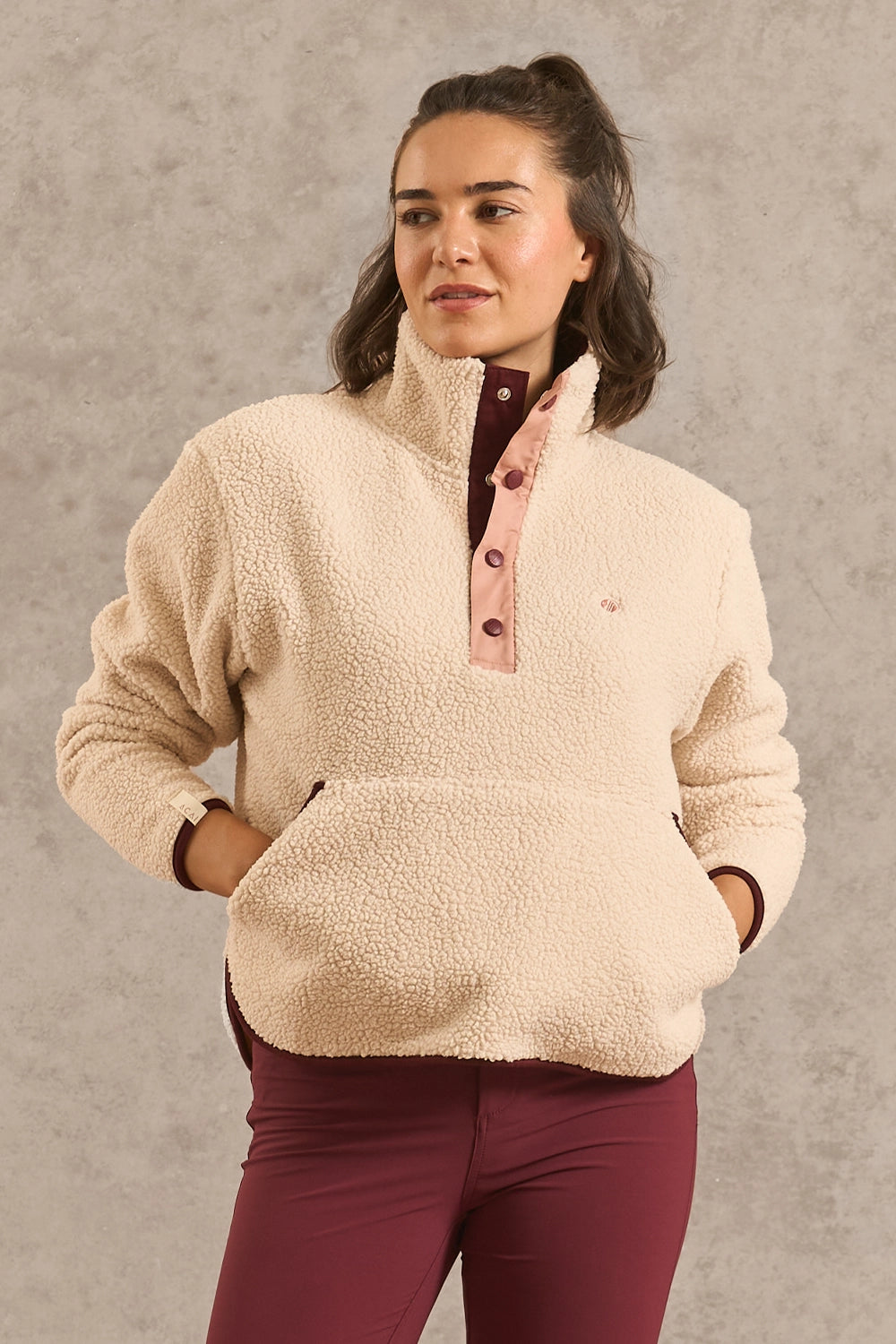 A woman wearing a cream sherpa fleece quarter-zip pullover with a pink button placket and burgundy pants, standing against a textured gray wall.