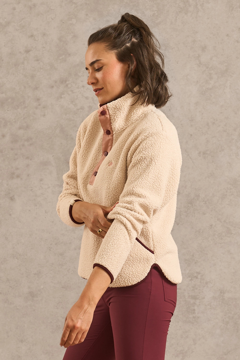 A woman wearing a cream sherpa fleece quarter-zip pullover jacket with dark brown trim, paired with burgundy leggings, standing against a textured gray wall.