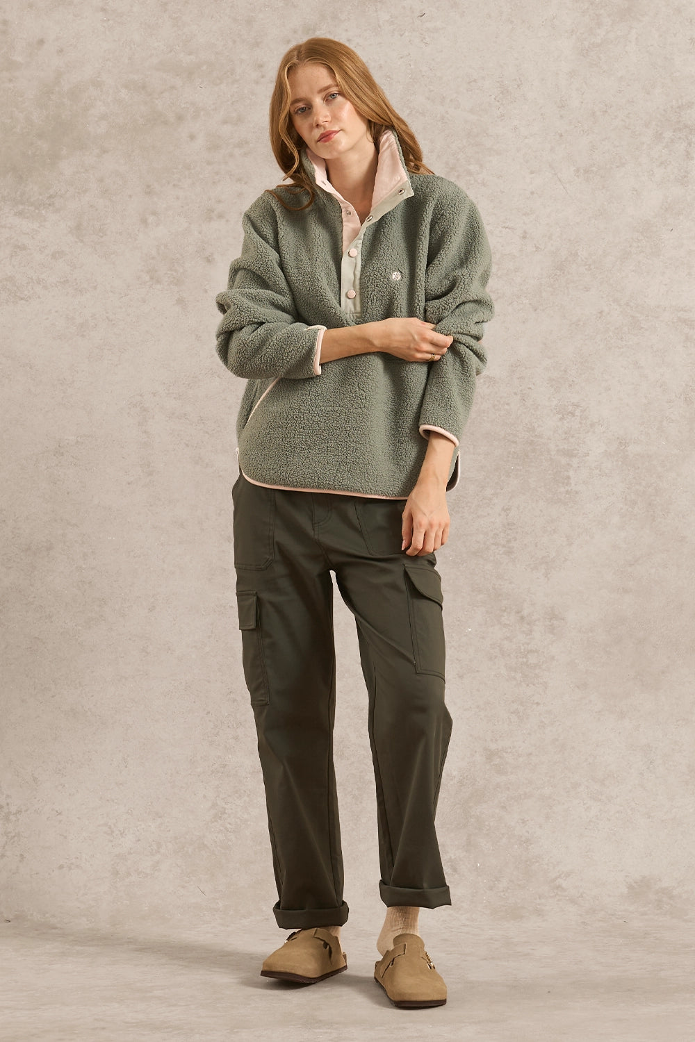 A woman stands against a textured light gray studio backdrop wearing a sage green sherpa fleece quarter-zip pullover with cream lining, dark olive cargo pants, and tan slip-on shoes.