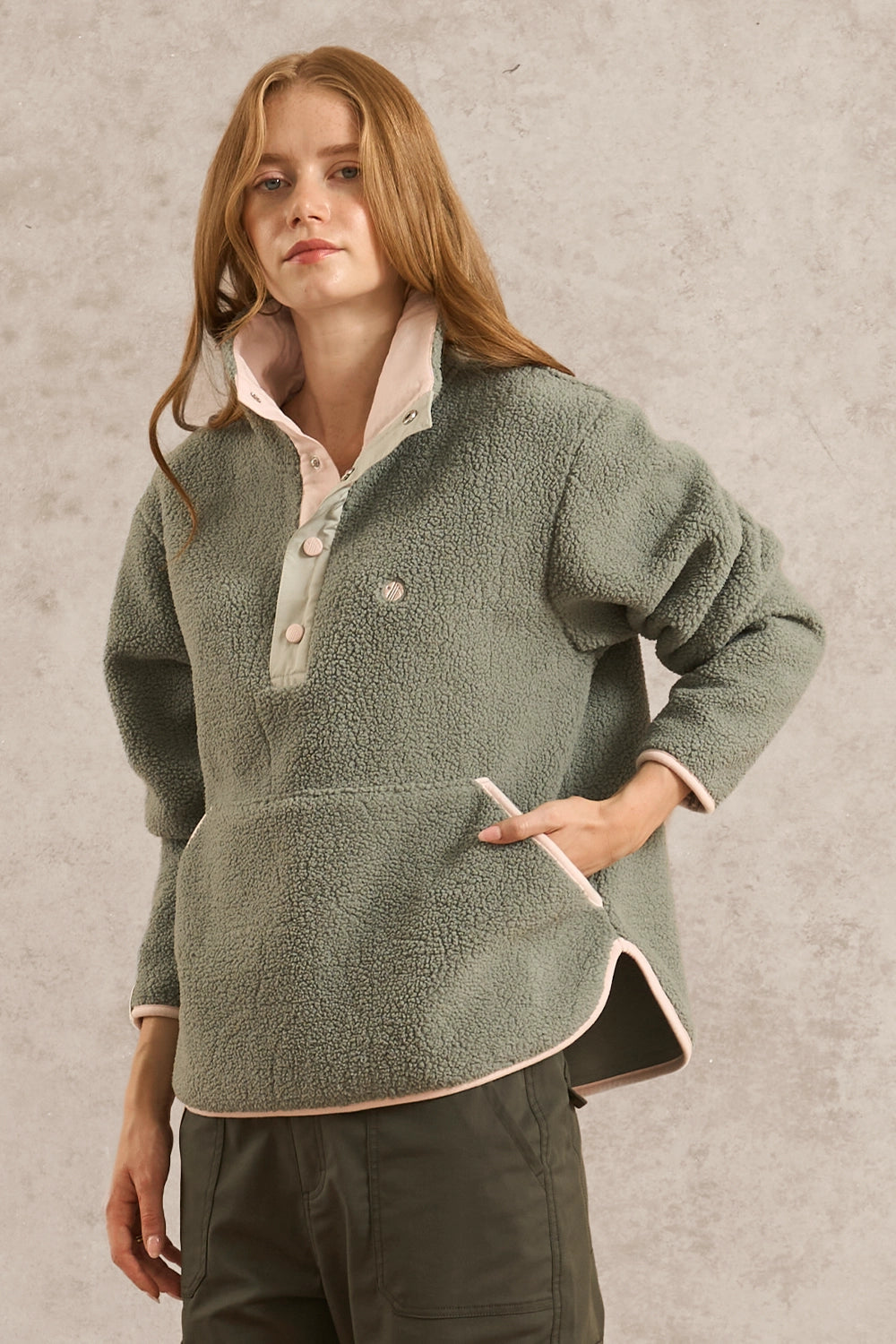 A woman wearing a sage green sherpa fleece pullover with light pink trim over a pale pink collared shirt, paired with dark olive pants, set against a textured light gray wall.