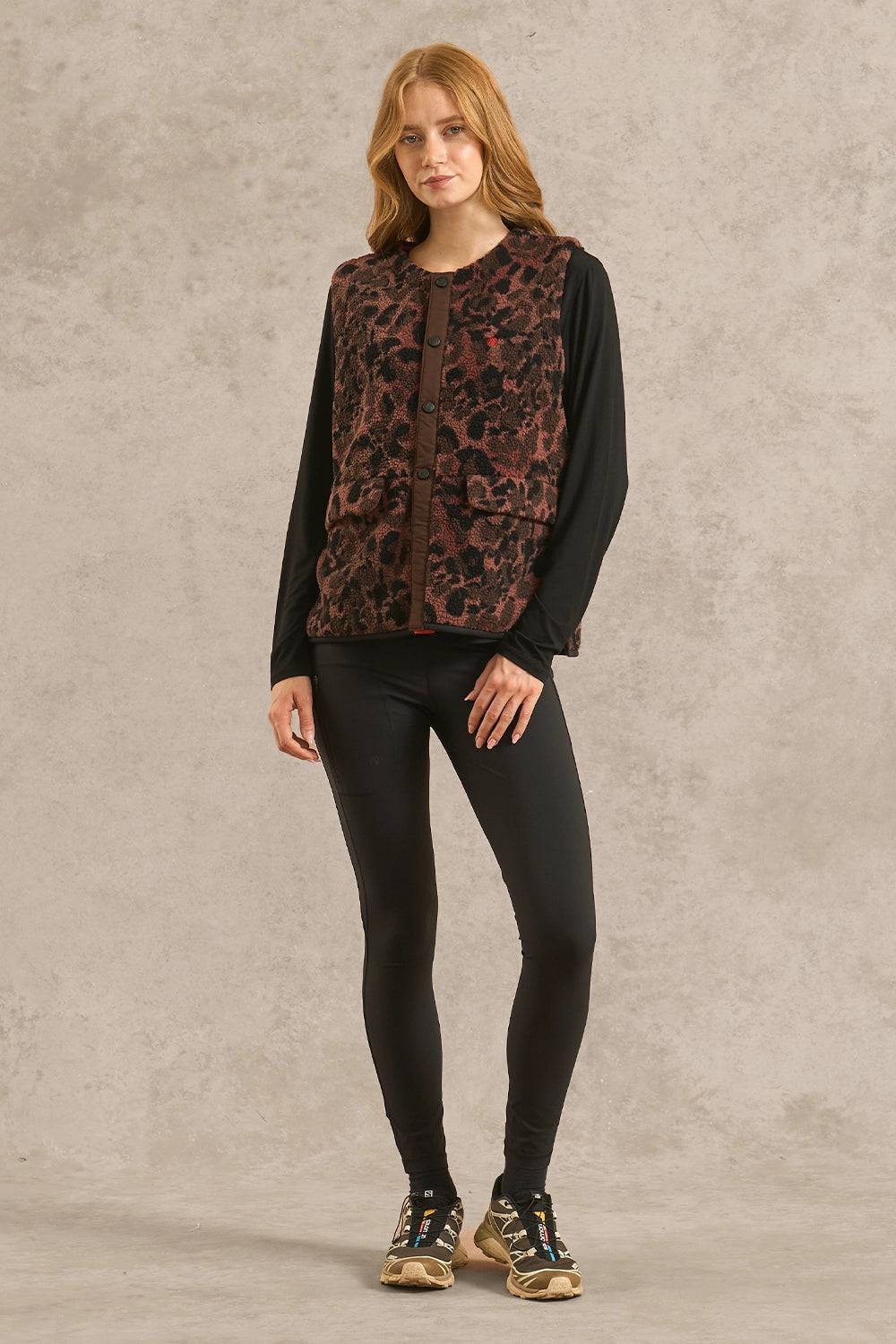 A woman modeling a sleeveless brown and black leopard-print vest over a black long-sleeve shirt, black leggings, and hiking shoes, standing against a textured light gray studio backdrop.