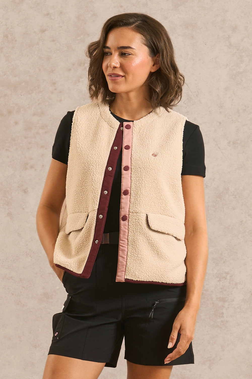 A woman modeling a cream sherpa fleece vest with burgundy and dusty pink trim over a black t-shirt and black shorts, set against a textured beige studio background.