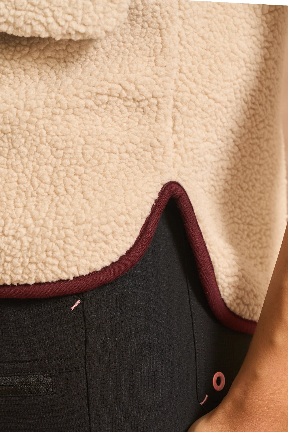 Close-up of a cream sherpa fleece garment with a dark burgundy trim, layered over black textured pants.