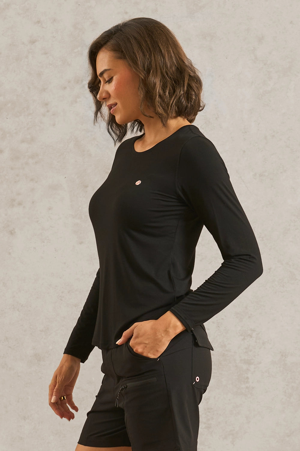 A woman wearing a black long-sleeve athletic top and matching black shorts, standing against a textured light gray wall.