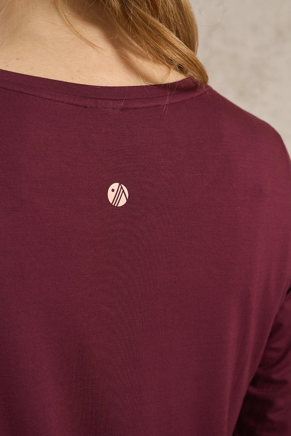 Close-up of a person wearing a burgundy long-sleeve top, showing the back of the neck and a small circular logo on the upper back.