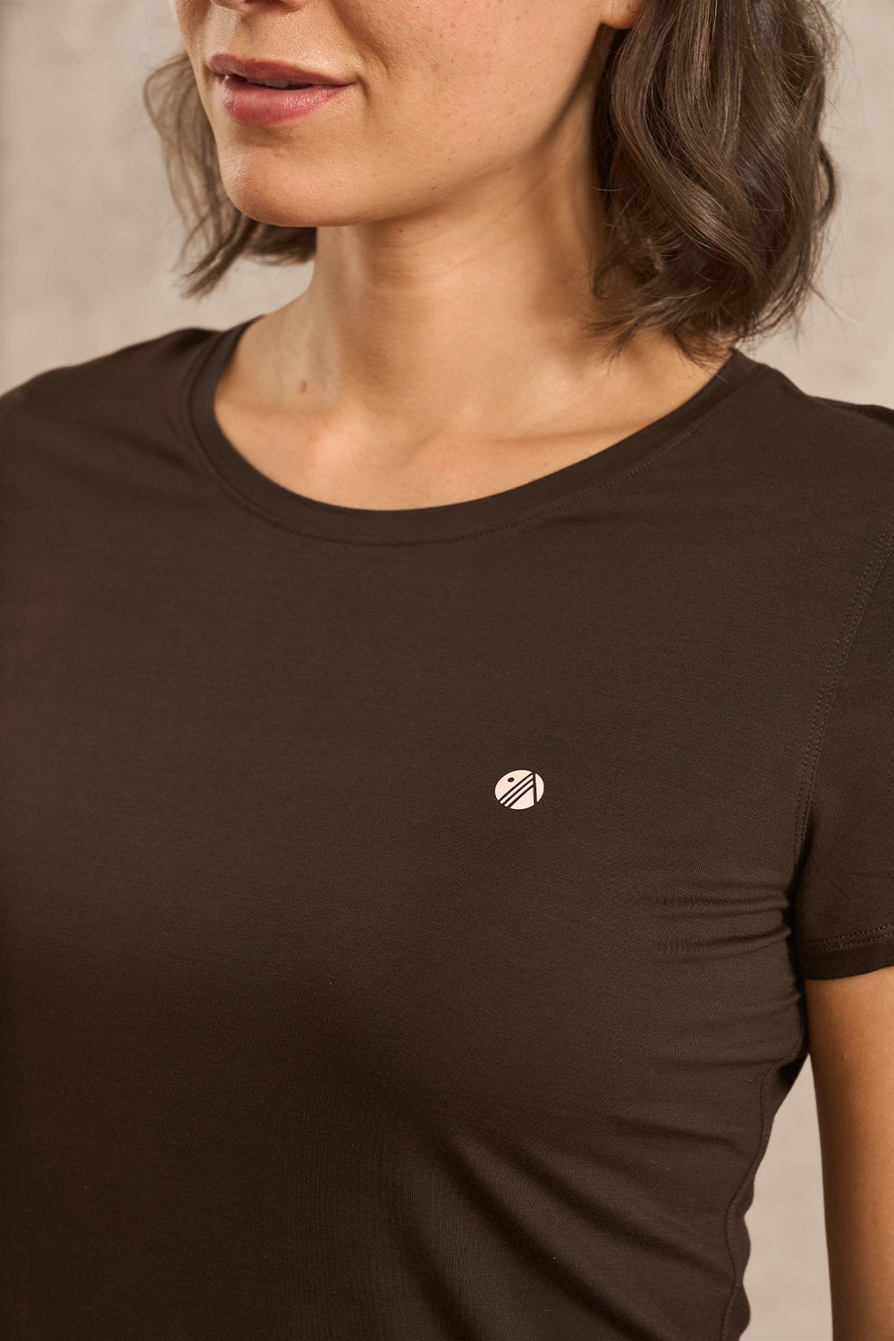 A close-up of a woman wearing a dark brown short-sleeve crewneck t-shirt with a small white circular logo on the left chest, set against a neutral beige background.