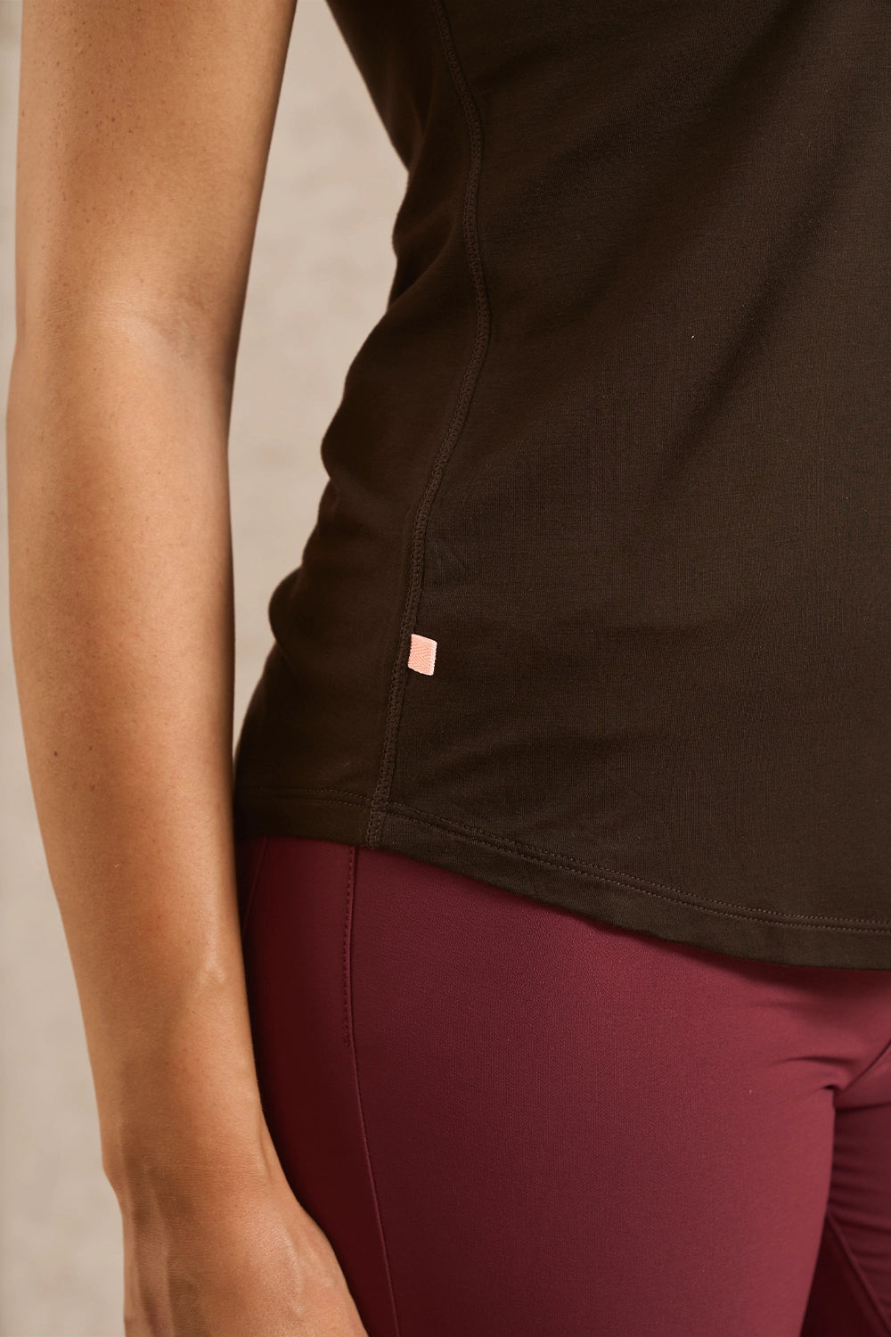 Close-up of a person wearing a dark brown athletic tank top and burgundy leggings, showing the side seam and a small pink brand tab.