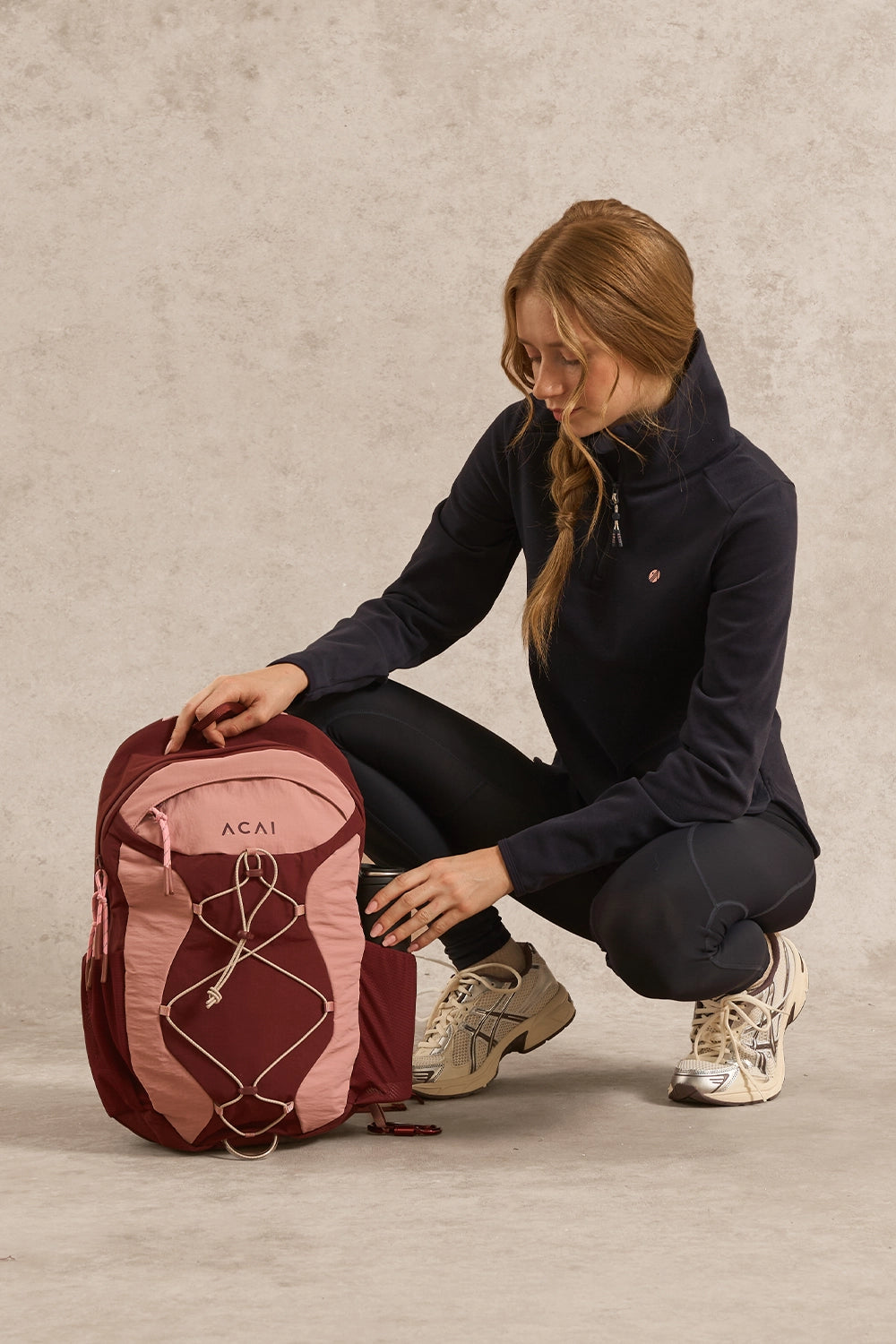 A woman crouches beside a pink and burgundy hiking backpack on a textured beige studio floor, wearing a navy fleece jacket, black leggings, and beige sneakers.