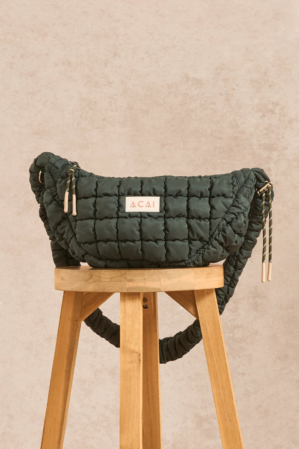 A dark green quilted crossbody bag with a central white label reading 'ACAI', displayed on a wooden stool against a textured beige background.