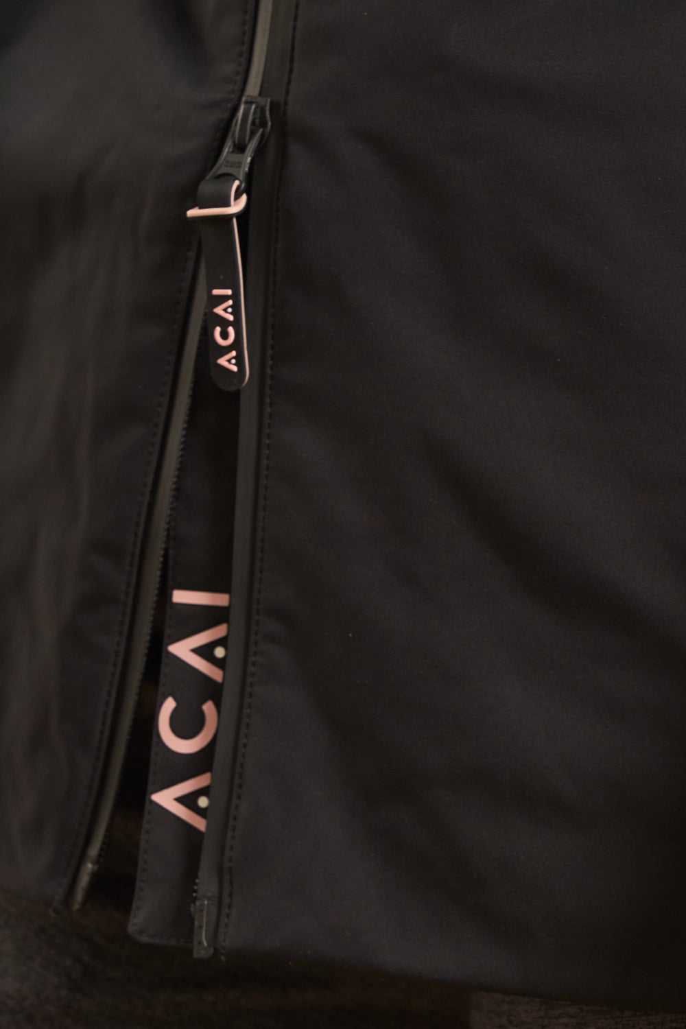 Close-up of a black zippered garment with a rose gold zipper pull and vertical ACAI branding printed in rose gold along the zipper track.