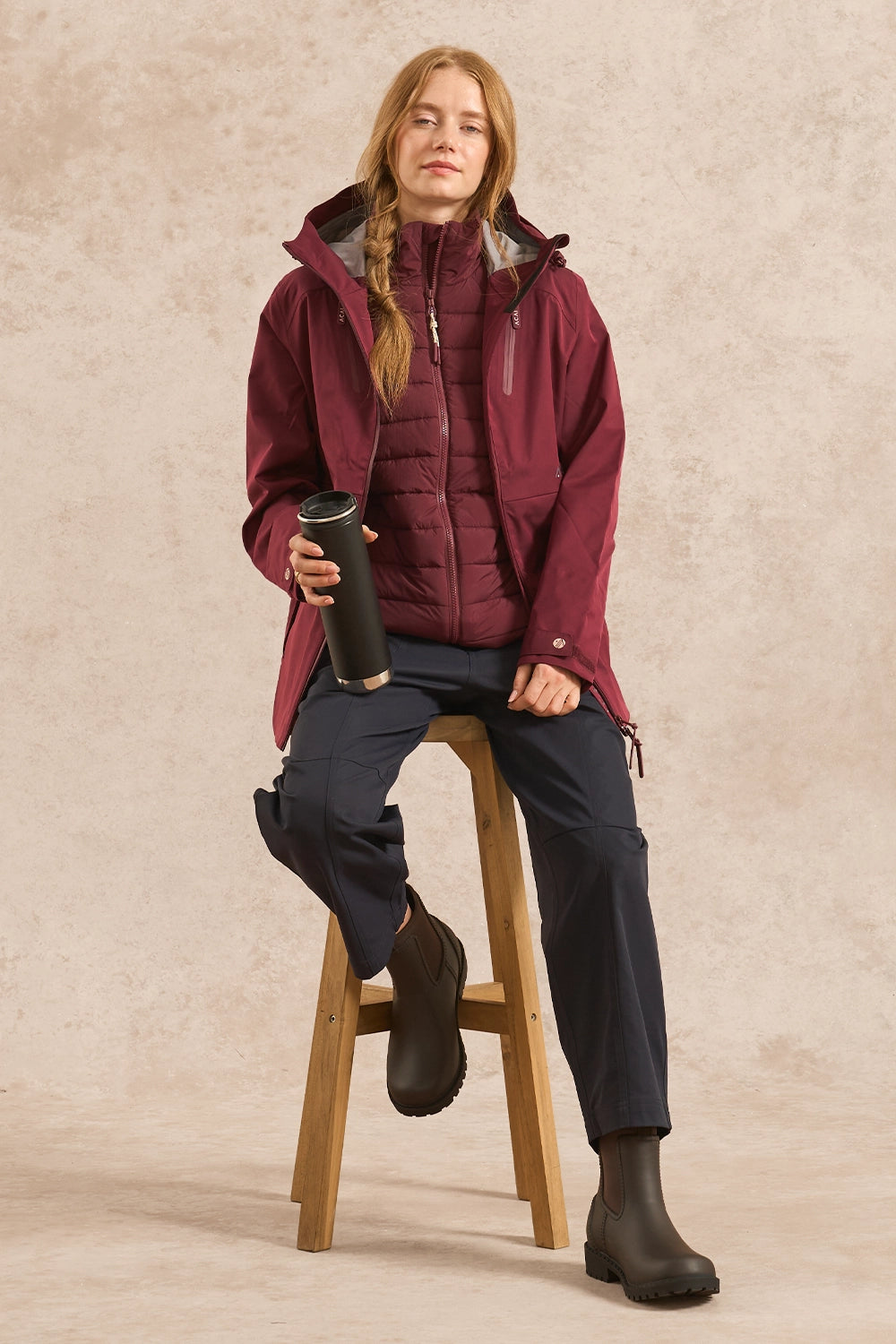 A woman sits on a wooden stool wearing a burgundy hooded puffer jacket over a matching quilted top, dark pants, and black boots, while holding a black travel tumbler against a textured beige studio backdrop.