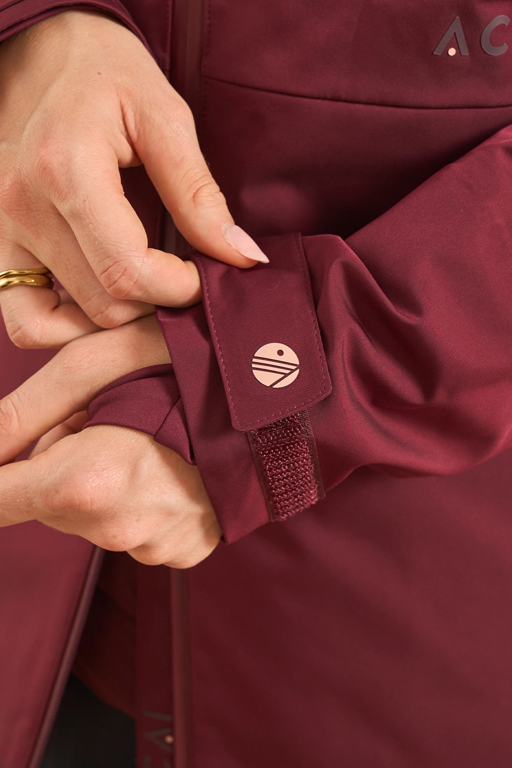 Close-up of a person's hands adjusting the cuff of a burgundy jacket, showing a small circular metallic logo patch and a hook-and-loop strap closure.