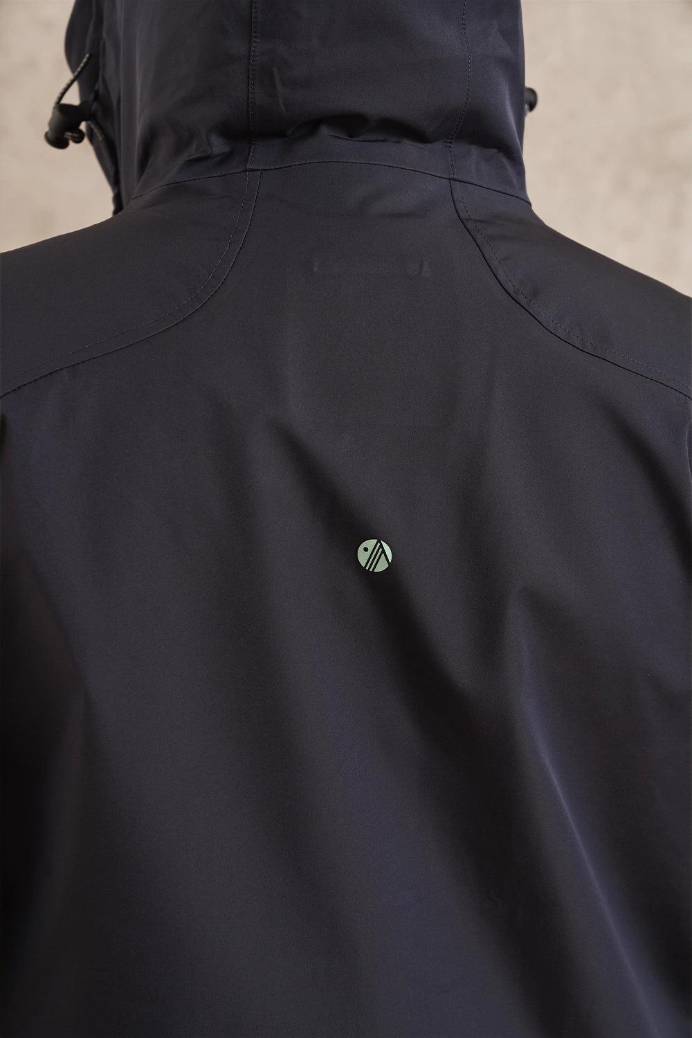 Close-up back view of a dark navy hooded jacket with a small circular green and white logo centered on the upper back.