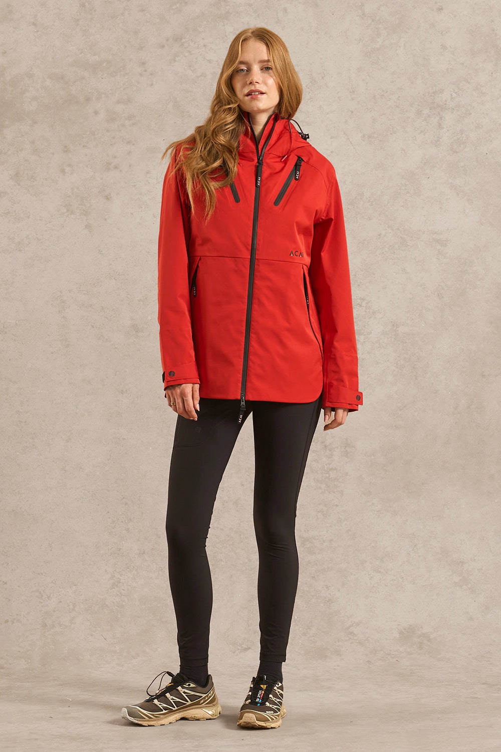 A woman modeling a bright red hooded outdoor jacket with black zippers, paired with black leggings and hiking shoes, standing against a textured light gray studio backdrop.