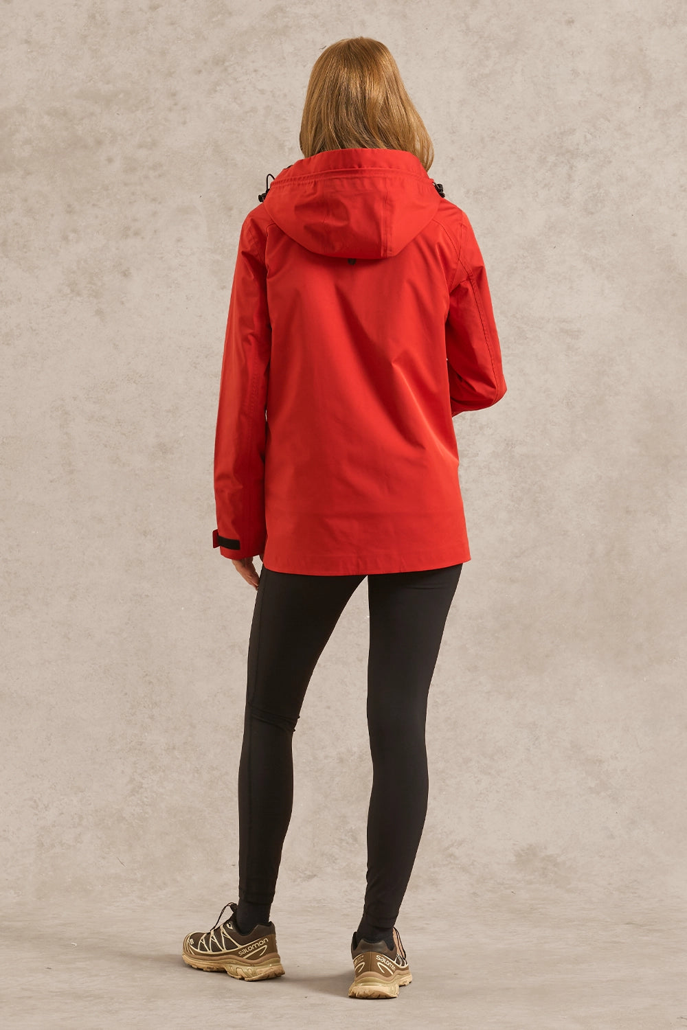 A woman viewed from behind wearing a bright red hooded jacket, black leggings, and tan sneakers, standing against a textured light beige studio wall.