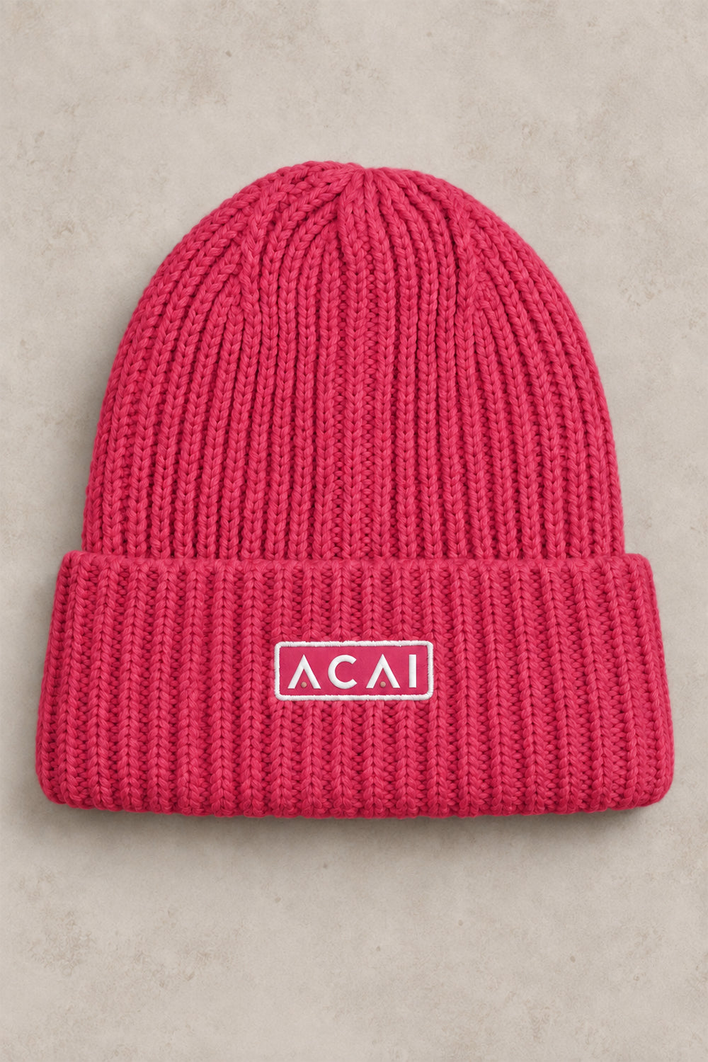 A bright pink ribbed knit beanie hat with a folded cuff, featuring a white rectangular patch with the text 'ACAI' in white on the front, set against a neutral textured gray background.