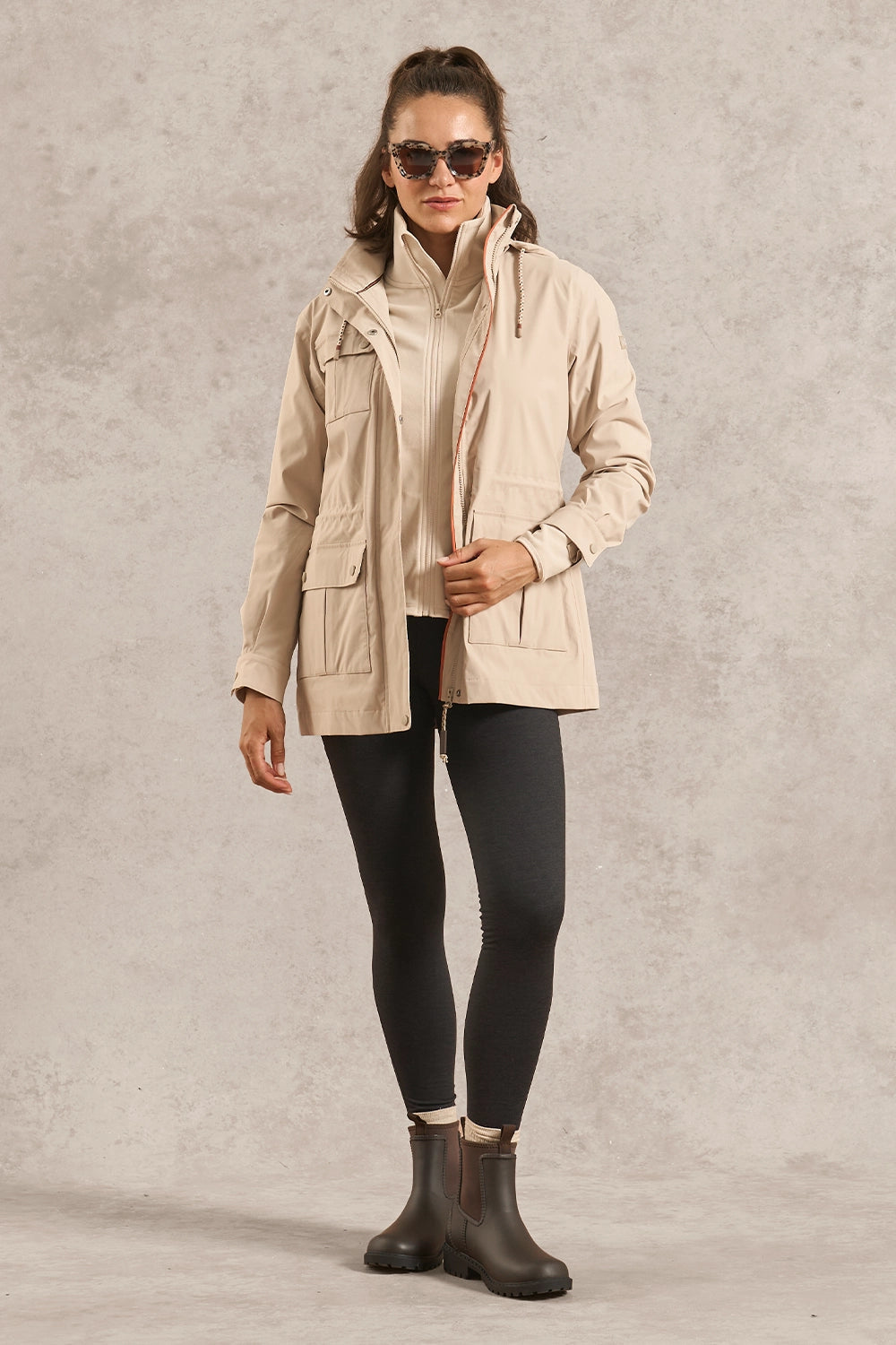 A woman modeling a light beige hooded jacket over a cream ribbed top, black leggings, and dark brown boots, standing against a textured light gray studio backdrop.