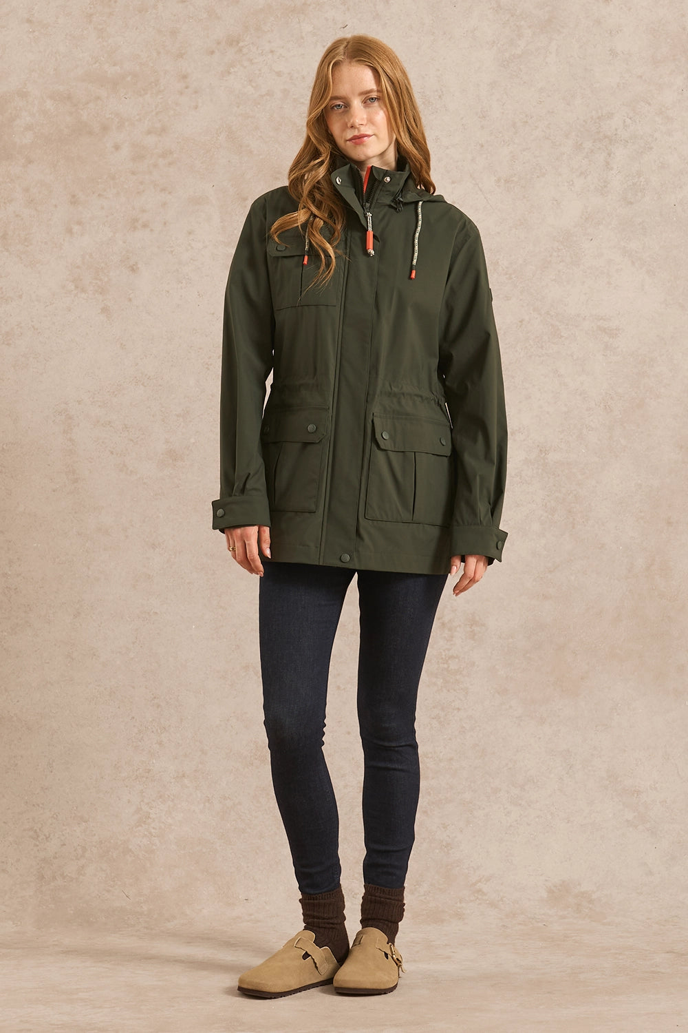 A woman modeling an olive green hooded utility jacket, paired with dark blue skinny jeans, brown socks, and tan moccasin-style shoes, standing against a textured beige studio backdrop.