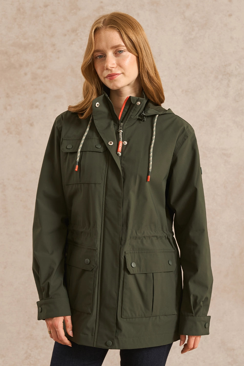 A woman wearing an olive green hooded utility jacket with a front zipper, drawstring hood, and multiple flap pockets, standing against a textured beige studio background.