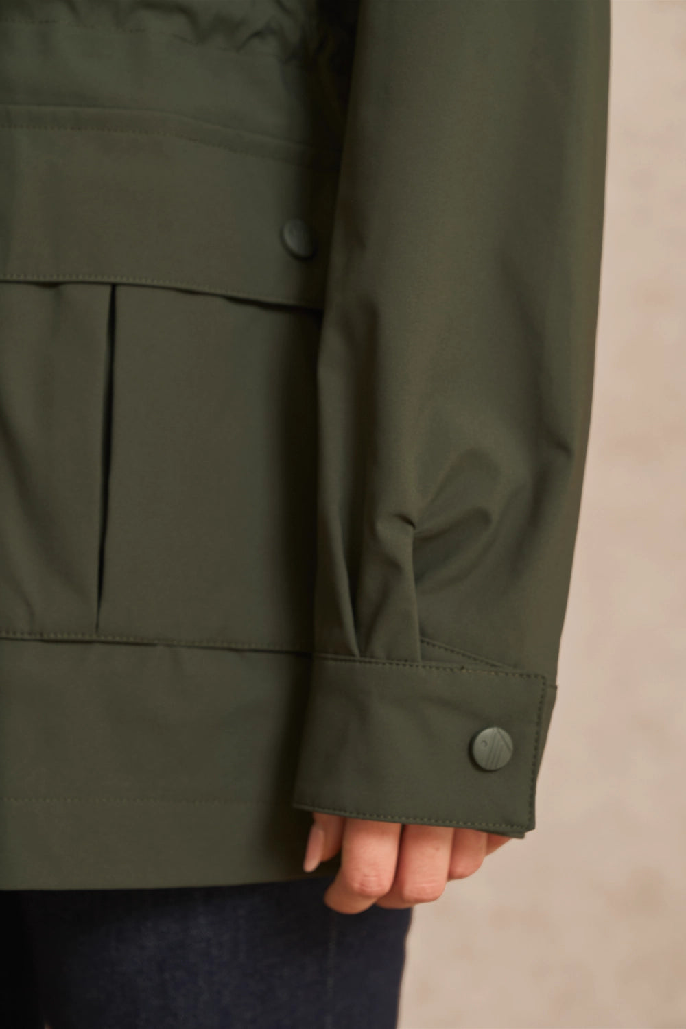 Close-up of a person wearing an olive green jacket, showing the sleeve, buttoned cuff, and part of the hand against a neutral beige background.