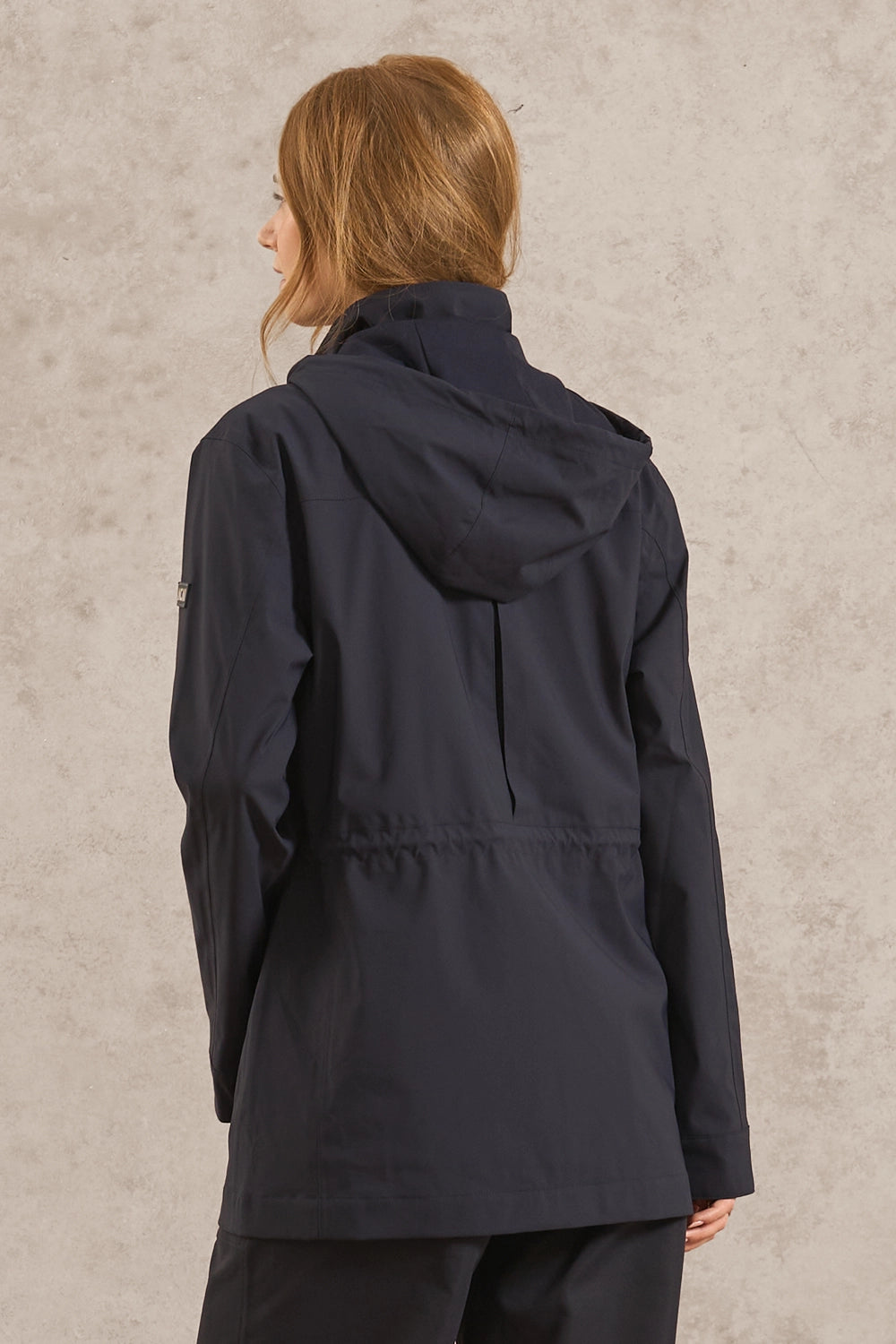 A woman viewed from the back wearing a dark navy hooded jacket and matching dark pants, standing against a textured light gray wall.