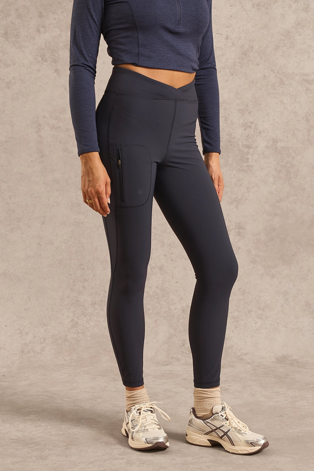 A model wearing high-waisted navy blue athletic leggings with a side zip pocket, paired with a navy long-sleeve crop top, beige socks, and white running shoes, standing against a textured gray studio backdrop.