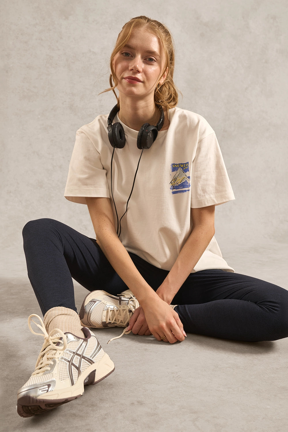 A young woman sits cross-legged on a textured gray floor, wearing a cream-colored graphic t-shirt, dark navy leggings, and cream sneakers with headphones around her neck.